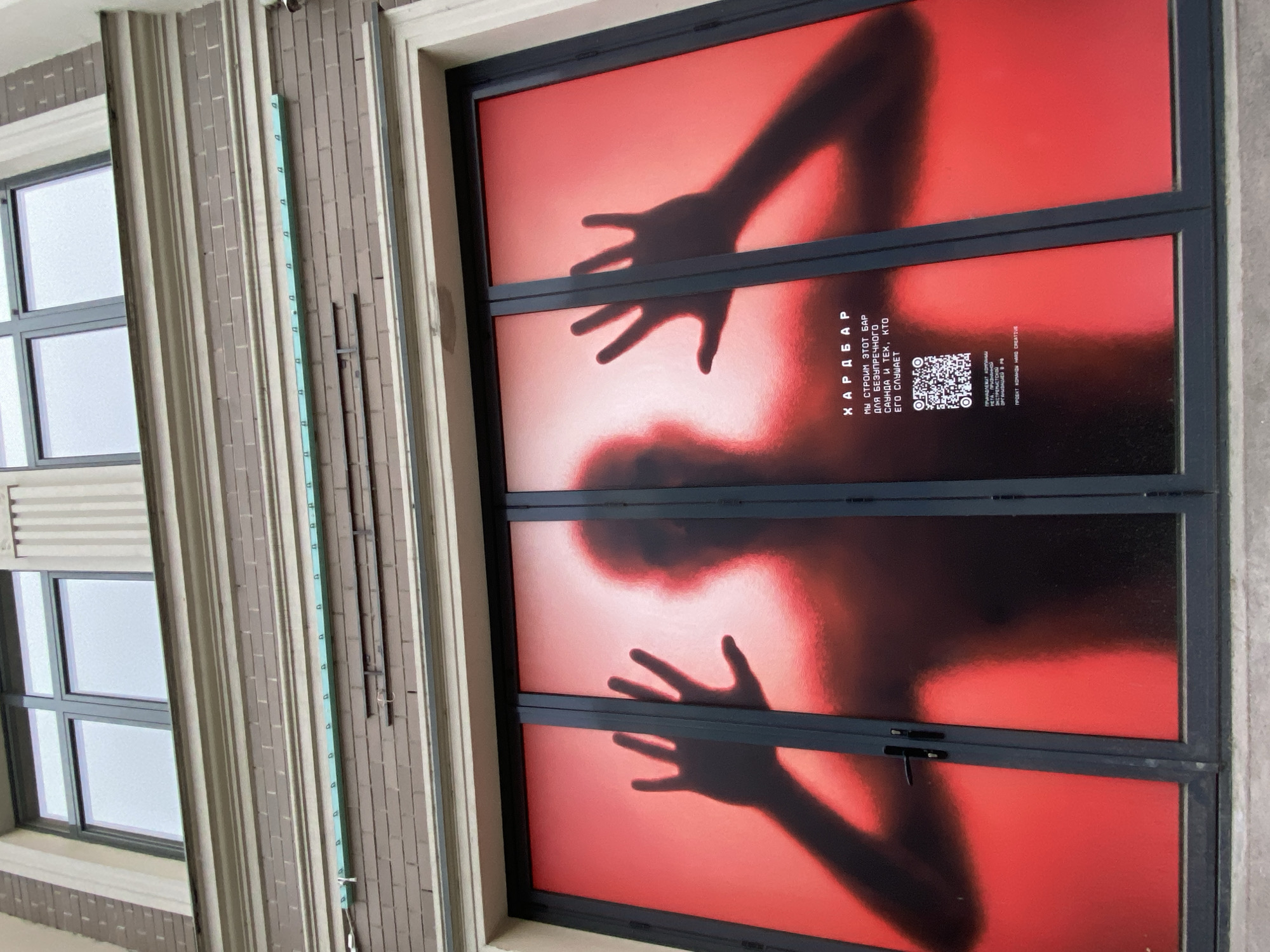 A close-up, eye-level shot depicts a poster or graphic on a window or door. The image is predominantly red, with a dark silhouette of a person with their hands outstretched. The poster or graphic is bisected by the frame of the window or door. The text "ХАРД БАР" and additional text including "МЫ СТРОИМ ЭТОТ БАР ДЛЯ БЕЗУПРЕЧНОГО САУНДА И ТЕХ, КТО ЕГО СЛУШАЕТ" and a QR code are visible. The location is Kazan, Russia.