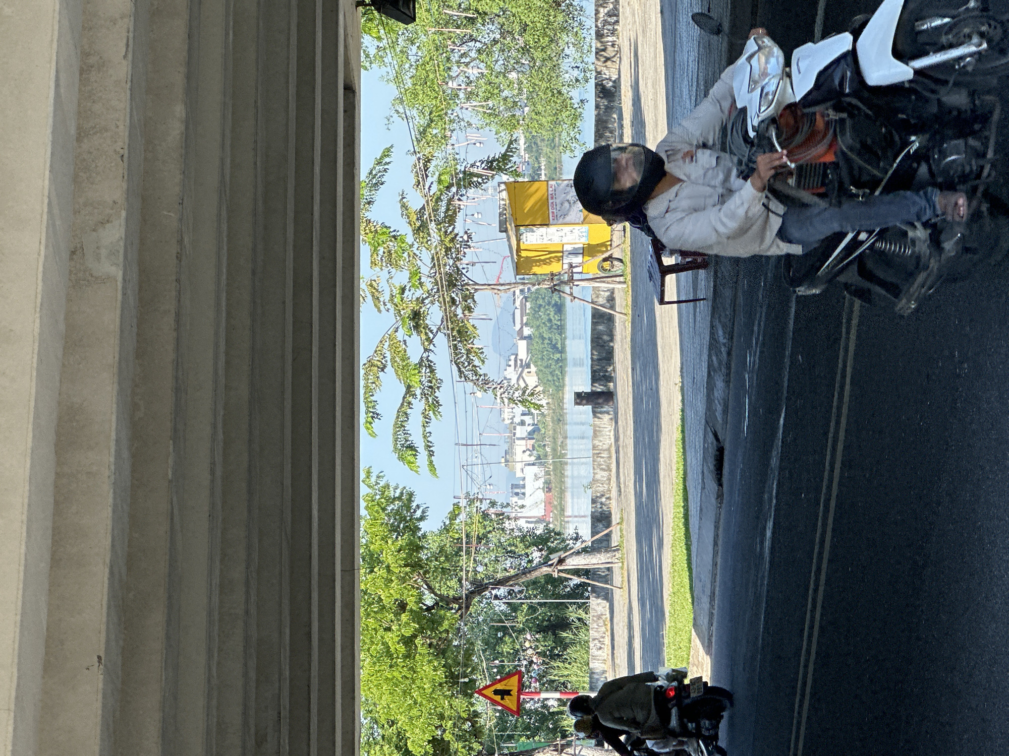 The image depicts a street scene in Da Nang, Vietnam, under an elevated concrete structure, likely a bridge or overpass. In the foreground, a person wearing a light-colored long-sleeved shirt, blue jeans, and a black helmet is riding a white and black motorcycle. The motorcycle has an orange accent visible beneath the rider. To the left, another motorcycle with two people wearing darker clothing is also on the road.
Beyond the road, a paved walkway lines a body of water, possibly a river or bay. Green trees and foliage are abundant on the other side of the walkway. A yellow kiosk with the words "KEM DUA MALAI" written on it, along with posters, is situated near the water's edge. A red traffic sign with a yellow triangle and a black pictogram of a pedestrian and a cyclist is visible on the left side of the scene. In the background, buildings and structures are visible across the water, and overhead, power lines and decorative lights hang from poles. The lighting suggests it is daytime, with sunlight casting shadows on the road and ground.