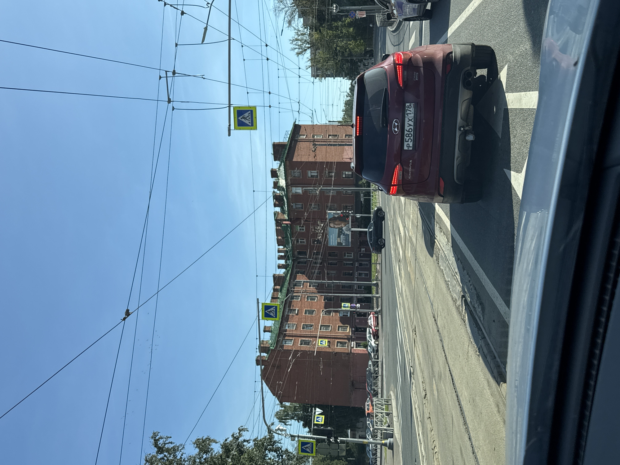The image captures a street scene in Saint Petersburg, Russia, on a clear day. A red Hyundai Santa Fe is in the foreground, facing away from the camera, with its license plate reading "P586YX 178 RUS". The car is stopped at a traffic light, in a lane marked with white arrows on the asphalt.
Behind the car, a large, multi-story brick building dominates the background, characterized by numerous windows and a distinctive green roofline with dormer-like structures. Several pedestrian crossing signs (yellow squares with a blue pedestrian symbol) are visible, suspended over the road by cables. Traffic lights are present, with one displaying red.
The street itself shows tram tracks embedded in the asphalt, indicating this is a route for public transportation. Other vehicles, including cars and potentially a tram, are visible in the distance. Trees line the sides of the street, and overhead electrical wires crisscross the sky. The sky is a clear, bright blue, with no clouds visible. The overall impression is of an urban environment in motion, with traffic and pedestrian infrastructure characteristic of a European city.
