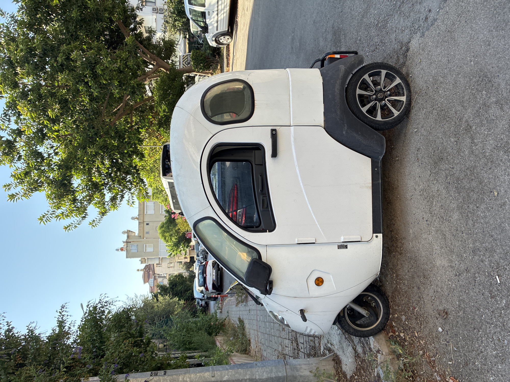 A white, three-wheeled, enclosed microcar is parked on a paved street next to a sidewalk lined with trees and buildings. The microcar has a rounded, bubble-like design with two side doors, a driver's side window, and a small oval window on the passenger side. It has black trim around the wheel wells and a black handle on the driver's side door. The vehicle is slightly dirty. In the background, there are other cars, trees, and multi-story buildings, typical of a city street in Alanya, Turkey. The sky is clear and blue, suggesting a sunny day.