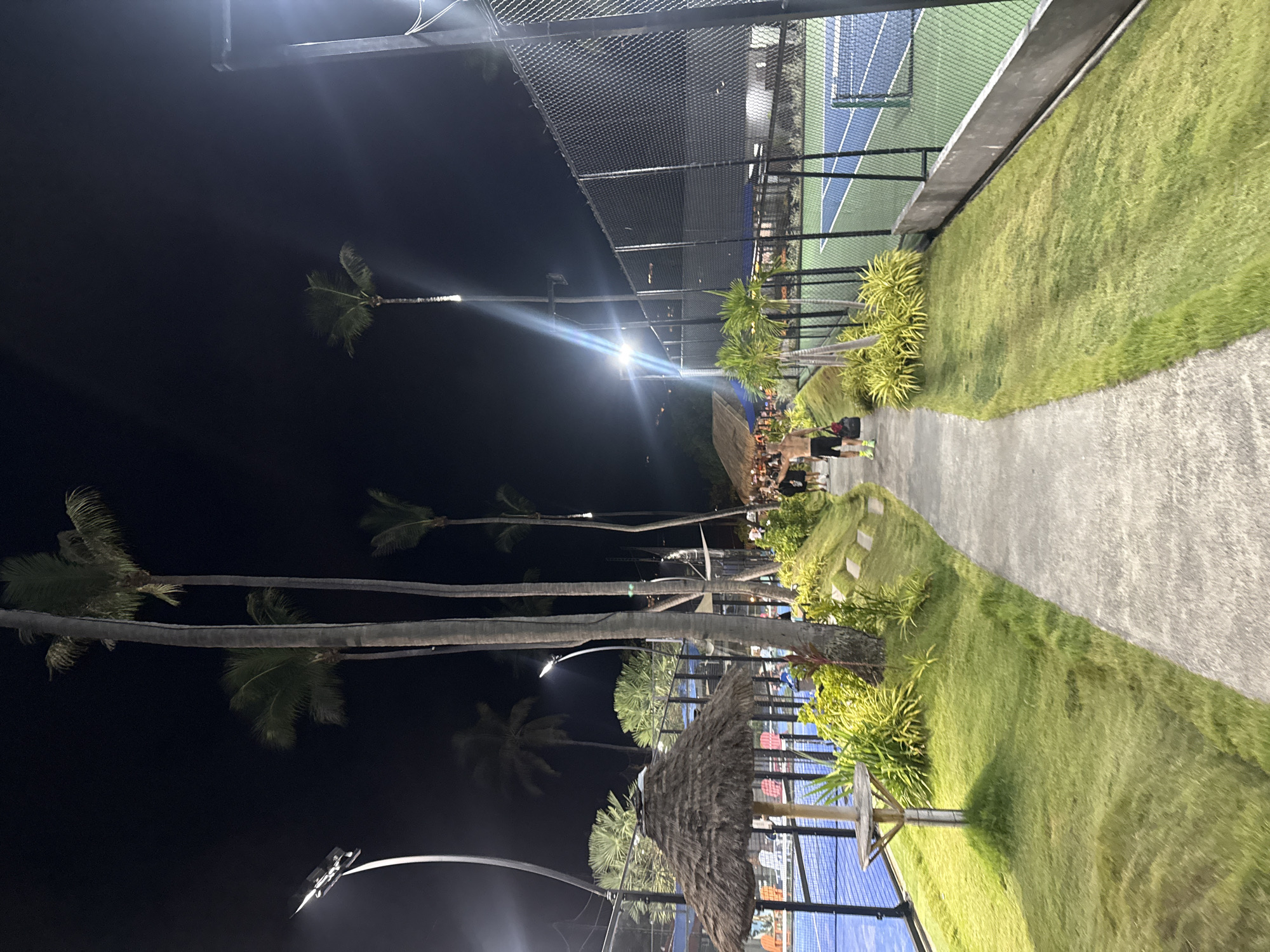 An outdoor sports complex in Ko Samui District, Thailand, features illuminated tennis courts at night. A paved walkway runs alongside a meticulously manicured lawn, bordered by a chain-link fence and lush vegetation, including palm trees and tropical plants. Several individuals are visible, with one man walking along the path, carrying a bag. In the background, parts of the blue tennis courts are visible, with netting and seating areas. The scene is lit by bright floodlights, casting long shadows and creating a dynamic atmosphere. A thatched-roof structure, possibly a shelter or bar, is also present on the left side of the image.