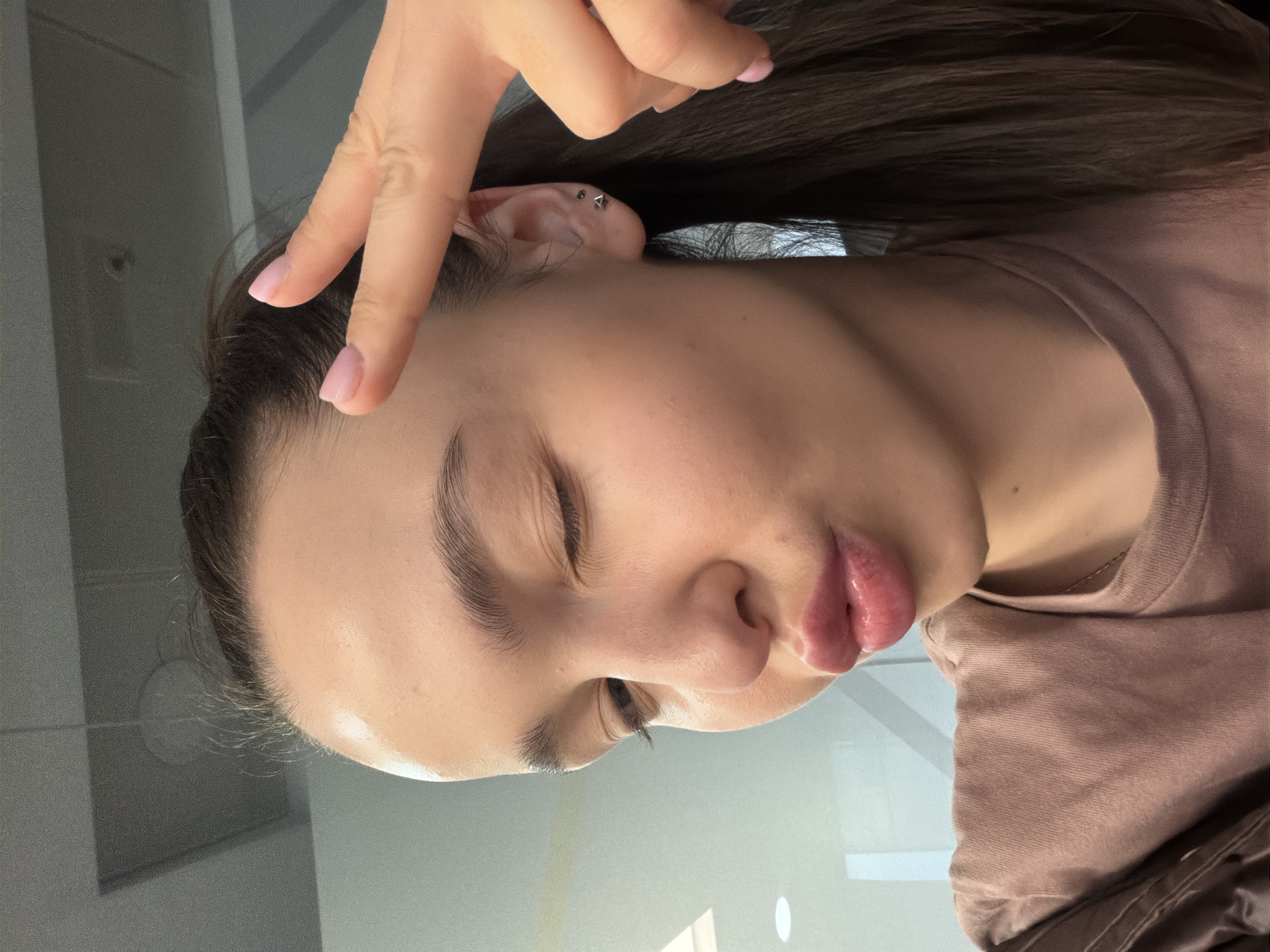 A close-up, selfie-style, slightly out-of-focus photograph depicts a woman from a low angle with her head tilted. She has fair skin, dark eyebrows, and full lips. Her eyes are mostly closed. Part of her dark hair is visible above and behind her head. A hand with manicured nails is raised, forming a 'V' shape with two fingers, near her hairline. An earring is visible in her ear. She is wearing a muted brown or mauve t-shirt. The background is a mix of neutral colors, possibly a wall or surface. The location cannot be determined from this image.