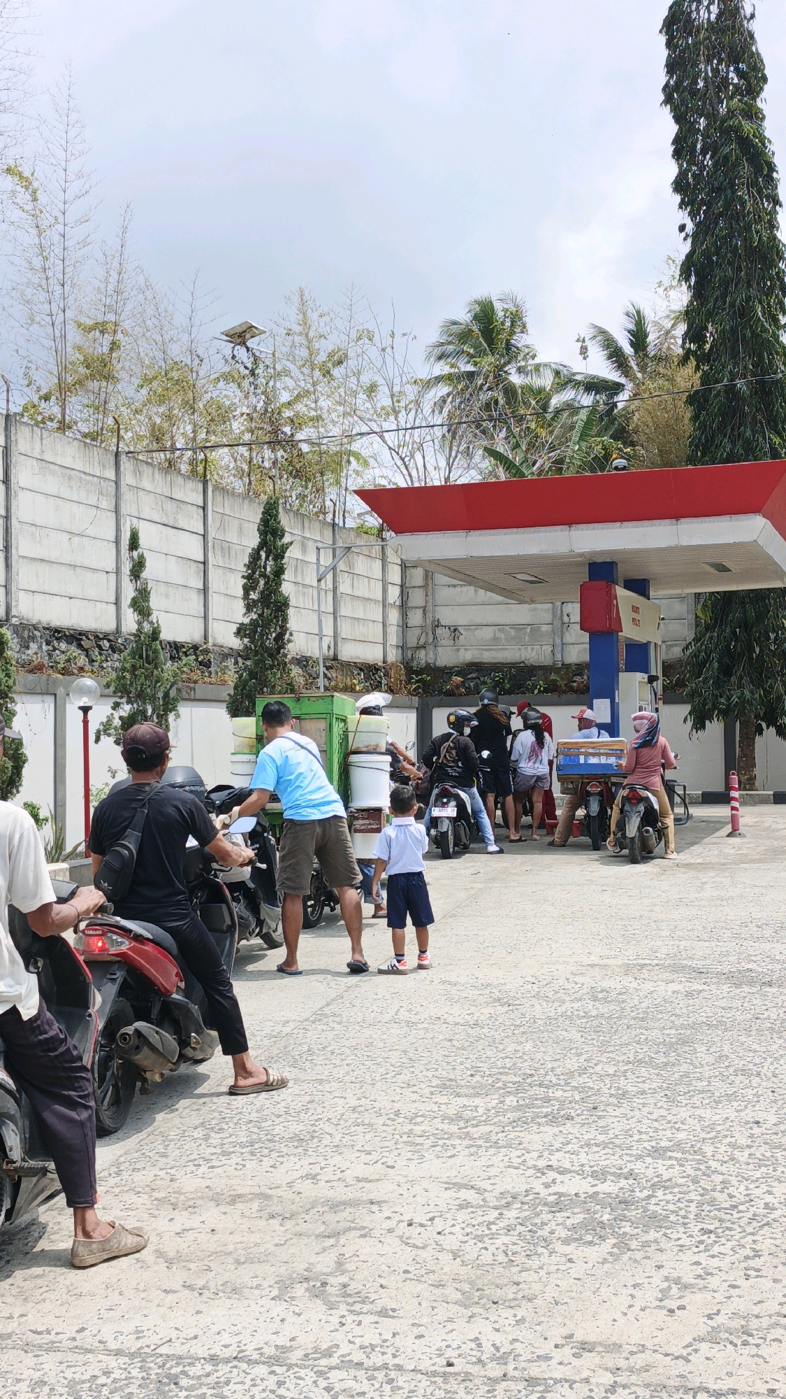 A group of people are gathered at a gas station in Sukaresik, Indonesia. The gas station has a red and white canopy over two fuel pumps. Several people are on motorcycles, some waiting in line and others attending to their vehicles. One man in a blue shirt appears to be interacting with a green cart stacked with white containers. A young boy in a white shirt and blue shorts stands near the cart. In the background, there is a concrete wall, trees, and a tall evergreen tree. The ground is paved and appears weathered. The sky is overcast.