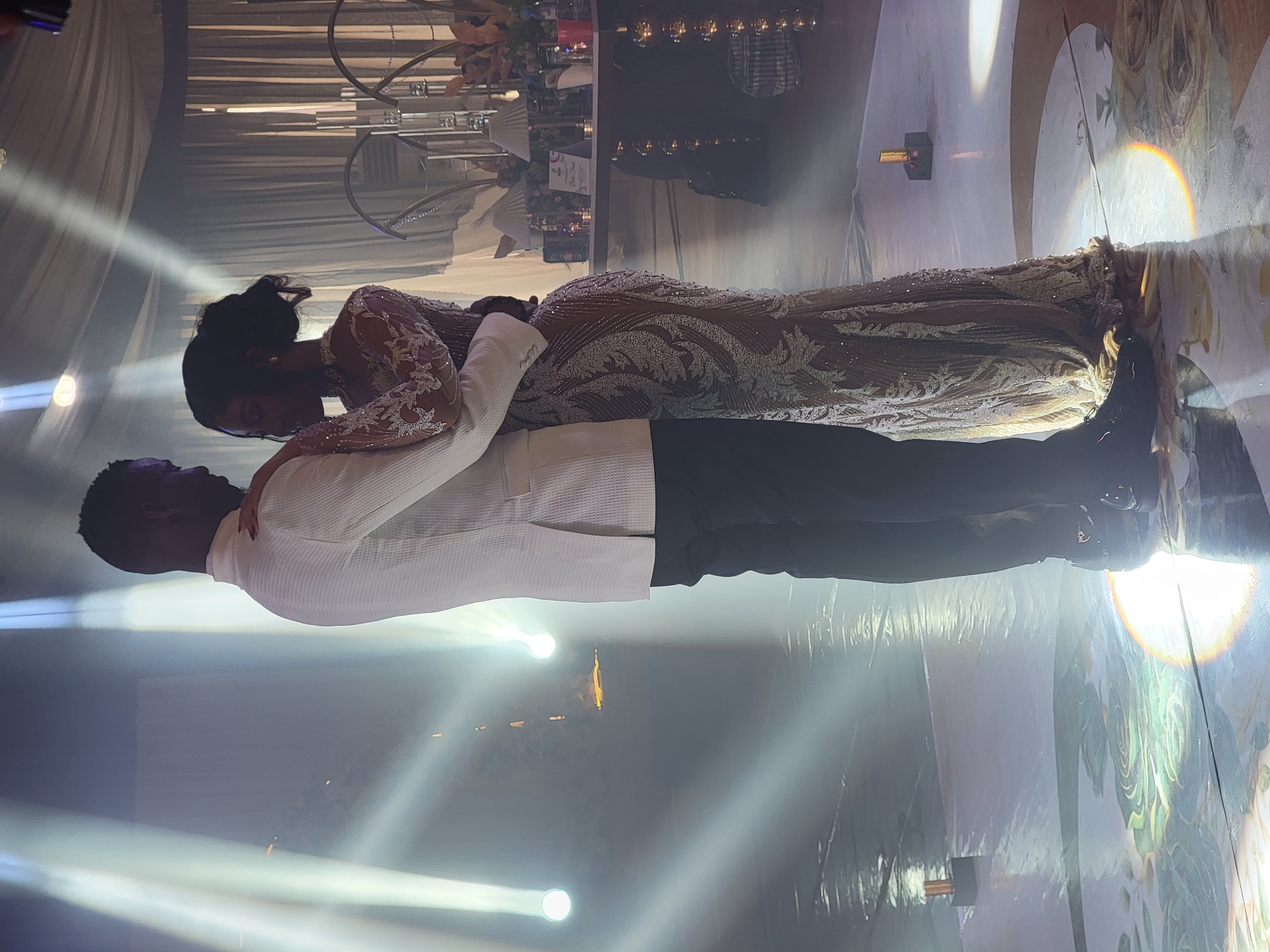 A couple slow dances intimately at what appears to be a formal indoor event, likely a wedding reception, in Barden Yana, Nigeria. The image is brightly lit by multiple spotlights, creating dramatic beams of light and lens flares across the hazy atmosphere.
The woman, with her head resting gently on the man's shoulder, is wearing a long, elaborate gown adorned with intricate silver and white embroidery or beading, featuring a sheer long-sleeved design and a fitted silhouette. Her hair is styled up. The man is dressed in a crisp white collared jacket or shirt with subtle vertical textures, paired with dark trousers. He holds her close, with one arm around her waist.
The setting features a decorated floor with elegant white and gold floral patterns, including large rose designs, and some visible reflections of the lights. In the background, draped light-colored fabrics create a soft, elegant ambiance. Elevated decorative elements, possibly chandeliers or light fixtures, and floral arrangements are partially visible in the upper background, suggesting a sophisticated event venue. The strong artificial lighting indicates an evening or nighttime event, contributing to a romantic and celebratory mood. No visible text is present in the image.
