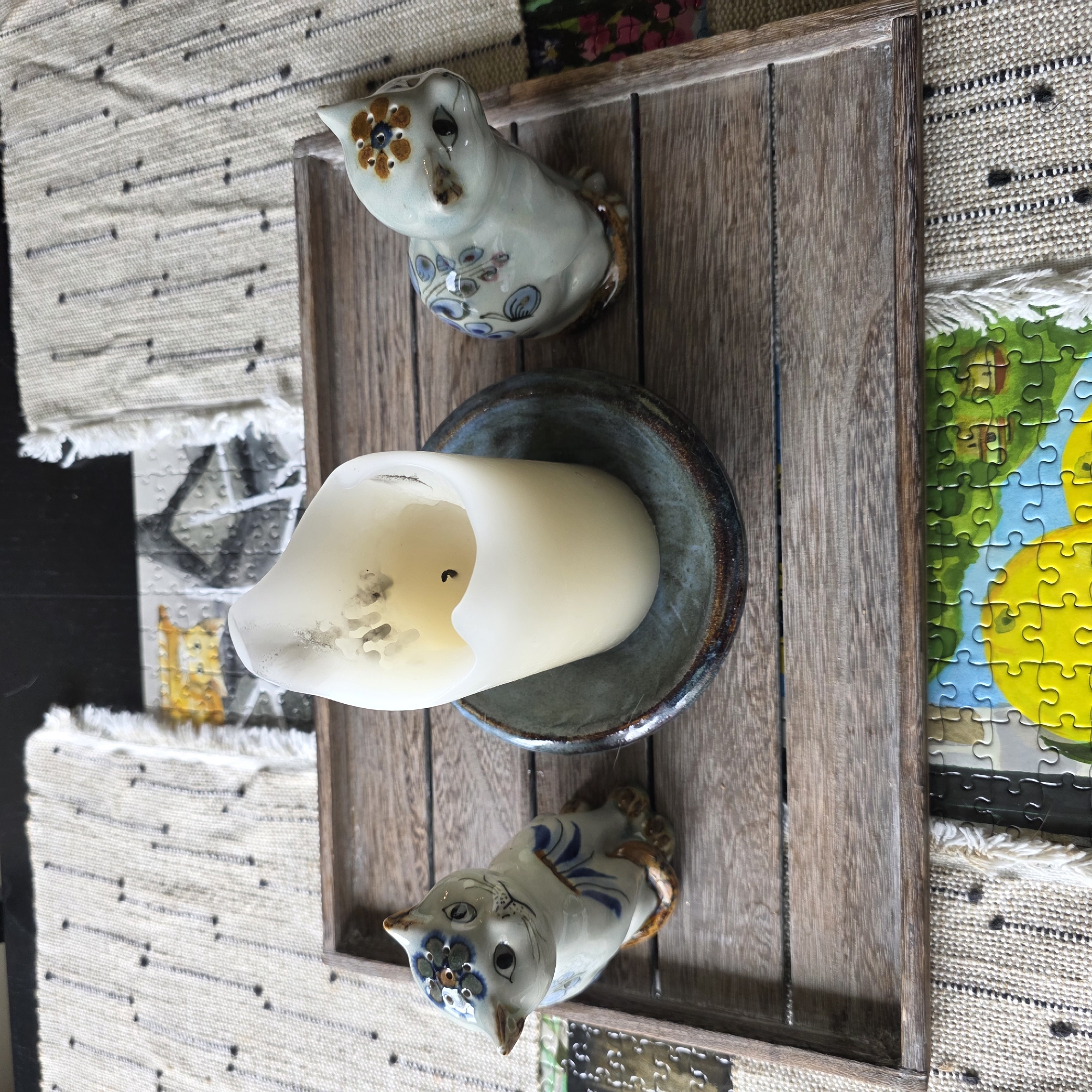 The image shows a rustic wooden tray on top of a cream-colored woven placemat with black stitch details. Inside the tray, there are two ceramic cat figurines and a thick, cream-colored pillar candle in a dark blue ceramic dish. The cat figurine on the left is facing forward and decorated with blue and white stripes and floral patterns. The cat figurine on the right is facing to the right and decorated with blue floral patterns and brown and orange flowers on its head. The candle is lit and has melted wax around its edges. The placemat has tasseled edges and is placed on a dark surface, possibly a table. In the background, part of a jigsaw puzzle with a colorful nature scene is visible. The scene is indoors, and the lighting suggests it could be daytime with natural light. The image is taken from a slightly overhead perspective. The location of this scene is unknown and cannot be determined from the image.