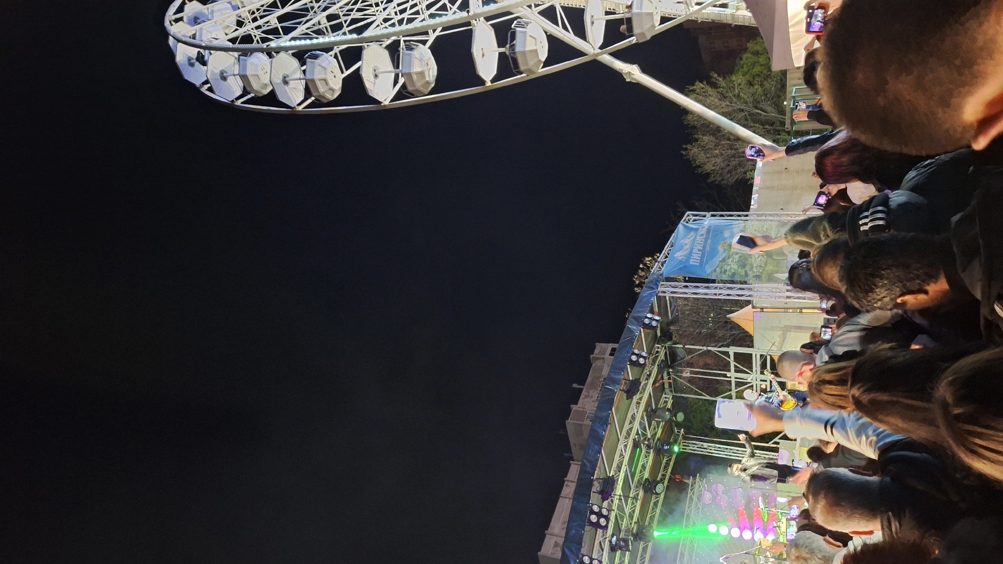 The image shows a night scene in Plovdiv, Bulgaria. A large Ferris wheel is visible in the upper left corner. Below the Ferris wheel, a crowd of people is gathered, many holding up phones to record a concert or event taking place on a stage. The stage is illuminated with green, pink, and white lights. There is a banner with the word "ПИРИНСКО" (Pirinsko) in Cyrillic text. The environment is dark, and the overall scene suggests a festive atmosphere.