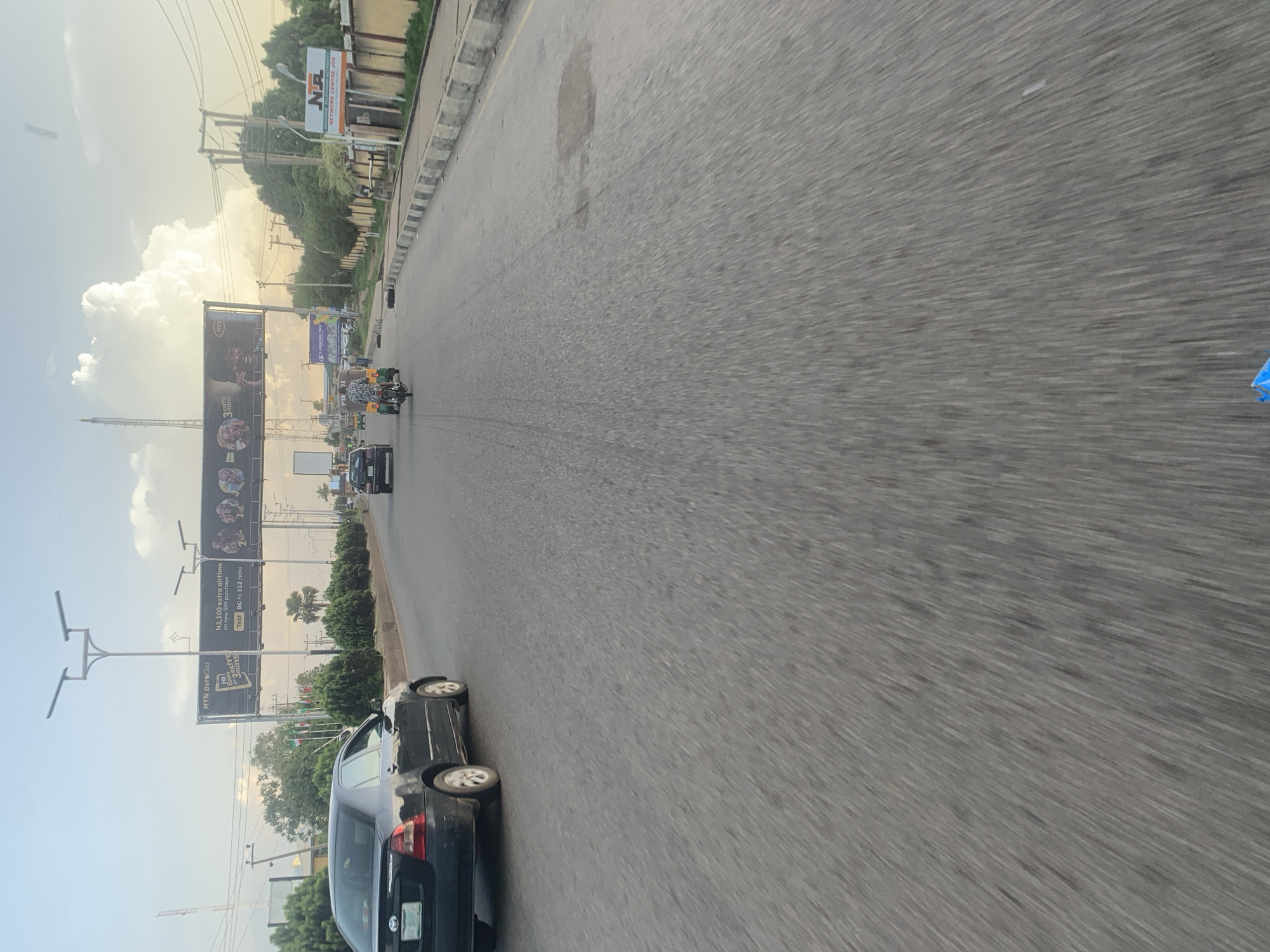 The image depicts a street scene in Jos, Nigeria, under a partly cloudy sky with the sun low on the horizon, suggesting either early morning or late afternoon. The road appears to be asphalt with a visible yellow line marking the lane. In the foreground, the road surface shows some discoloration, possibly from oil or water stains.
On the left side of the road, a black Toyota sedan is parked or moving slowly. Further down the road, another car, appearing darker in color, is visible. Dominating the background is a large billboard with advertisements. One prominent advertisement features the MTN logo and text, with images of people. Other advertisements, including one for "Long Long Tire," are also present.
Several streetlights with multiple lamps are positioned along the road. A few palm trees and dense green bushes line the roadside, separating the road from what appears to be commercial or residential buildings. On the right side of the road, a building with a yellow facade and a striped beige and brown wall is visible. A sign displaying "NA NETWORK CENTRE JOS" indicates the presence of a business.
Several people are visible. One person is riding a motorized tricycle (keke napep), and another individual is seen walking in the distance. The general atmosphere suggests a busy urban environment, common in a city like Jos. The motion blur in the image indicates it was likely taken from a moving vehicle.