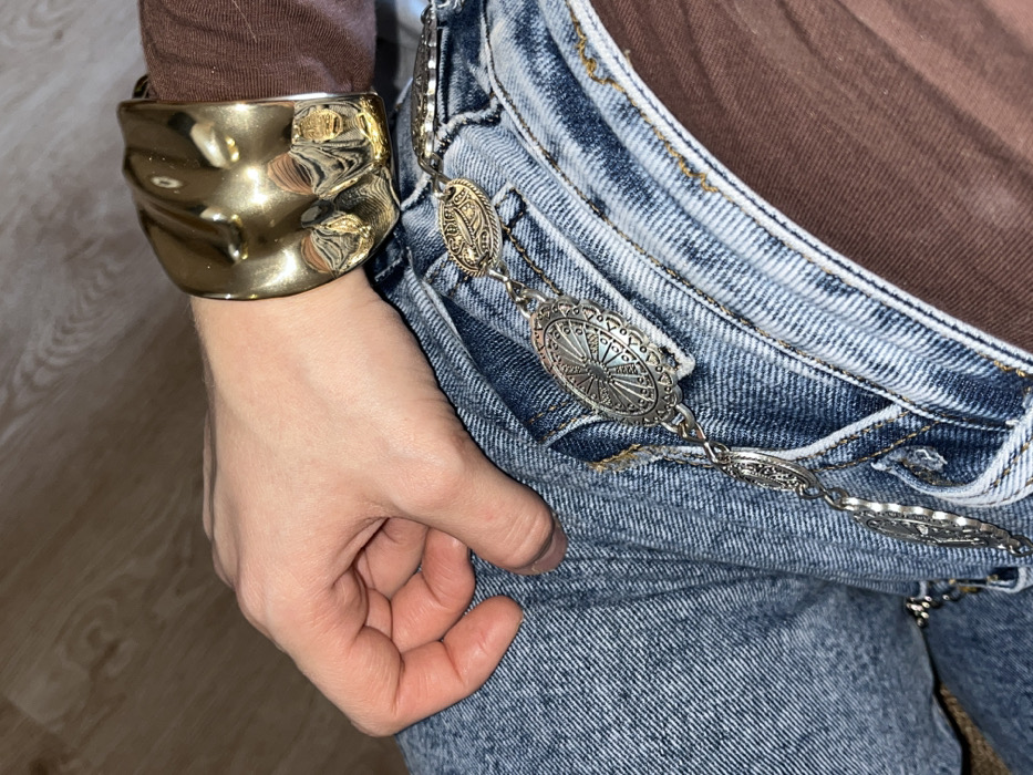 The image displays a close-up, downward-angled perspective of a human hand and a portion of the lower body, located in Zvezda, Russia. A person's bare left hand, exhibiting a light skin tone, is positioned with fingers slightly curled, resting atop an upper thigh. The wrist of this hand is adorned with a large, golden-hued, metallic cuff bracelet featuring an irregular, textured, and highly reflective surface. A brown fabric, consistent with a long-sleeved garment, is visible extending from beneath the cuff and on the right side of the frame. The lower body is encased in light-wash blue denim jeans, characterized by visible seams, pockets, and fabric texture. Draped across the jeans, from the top right towards the bottom left, is a silver-toned metal chain belt composed of multiple interconnected oval medallions, each featuring intricate embossed patterns and designs. The background to the upper left appears as a blurred light brown, possibly wood grain, surface.