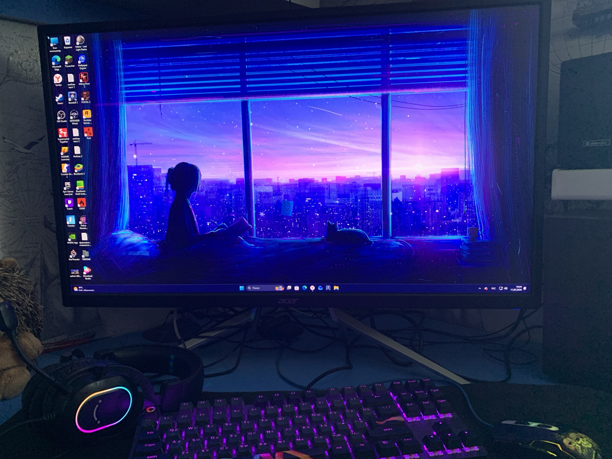 The image shows a computer monitor displaying a stylized image of a girl looking out a window at a cityscape with a sunset. The monitor, an Acer model, sits on a desk cluttered with wires. A keyboard with purple backlighting is in the foreground, along with a gaming mouse and a pair of headphones. The desktop on the monitor is filled with various game icons and software. The computer's system tray indicates a date of 11.05.2026. The general setting appears to be a room with a gaming setup. There is no indication of this image being in Severodvinsk, Russia.