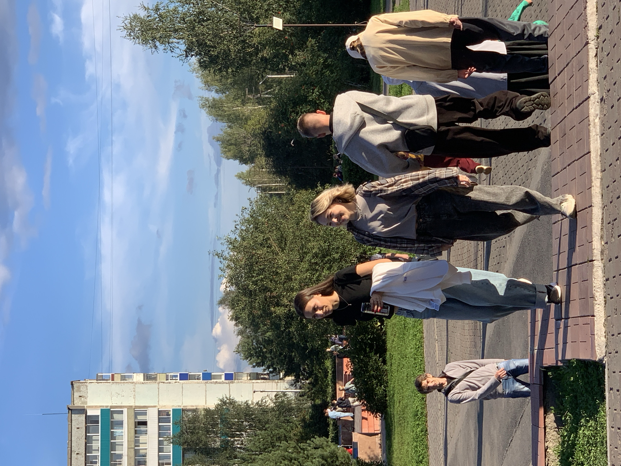 The image depicts an outdoor scene in Prokopyevsk, Russia, on a sunny day with a blue sky and scattered clouds. In the background, a multi-story residential building with light-colored facade and turquoise and blue accents is visible, along with trees. In the foreground, several people are walking on a paved walkway and a grassy area.
From left to right in the foreground:
A young man in a light gray hooded jacket and blue jeans stands facing the camera, with a dark bag slung across his chest. He is looking towards the camera.
To his right and further back, two women are walking away from the camera. The woman on the left has long dark hair and is wearing a black t-shirt and jeans, holding a white jacket and a can in her right hand. The woman on the right has blonde hair and is wearing a plaid shirt over a gray t-shirt and dark pants, with a necklace.
Further to the right, a person in a light gray hooded sweatshirt and dark pants is walking away from the camera, with a dark bag across their chest.
Behind them, a person in a light beige jacket and dark pants is also walking away from the camera, wearing a white cap.
Other people and a child in a green shirt are partially visible in the background. The environment includes green grass and paved surfaces.