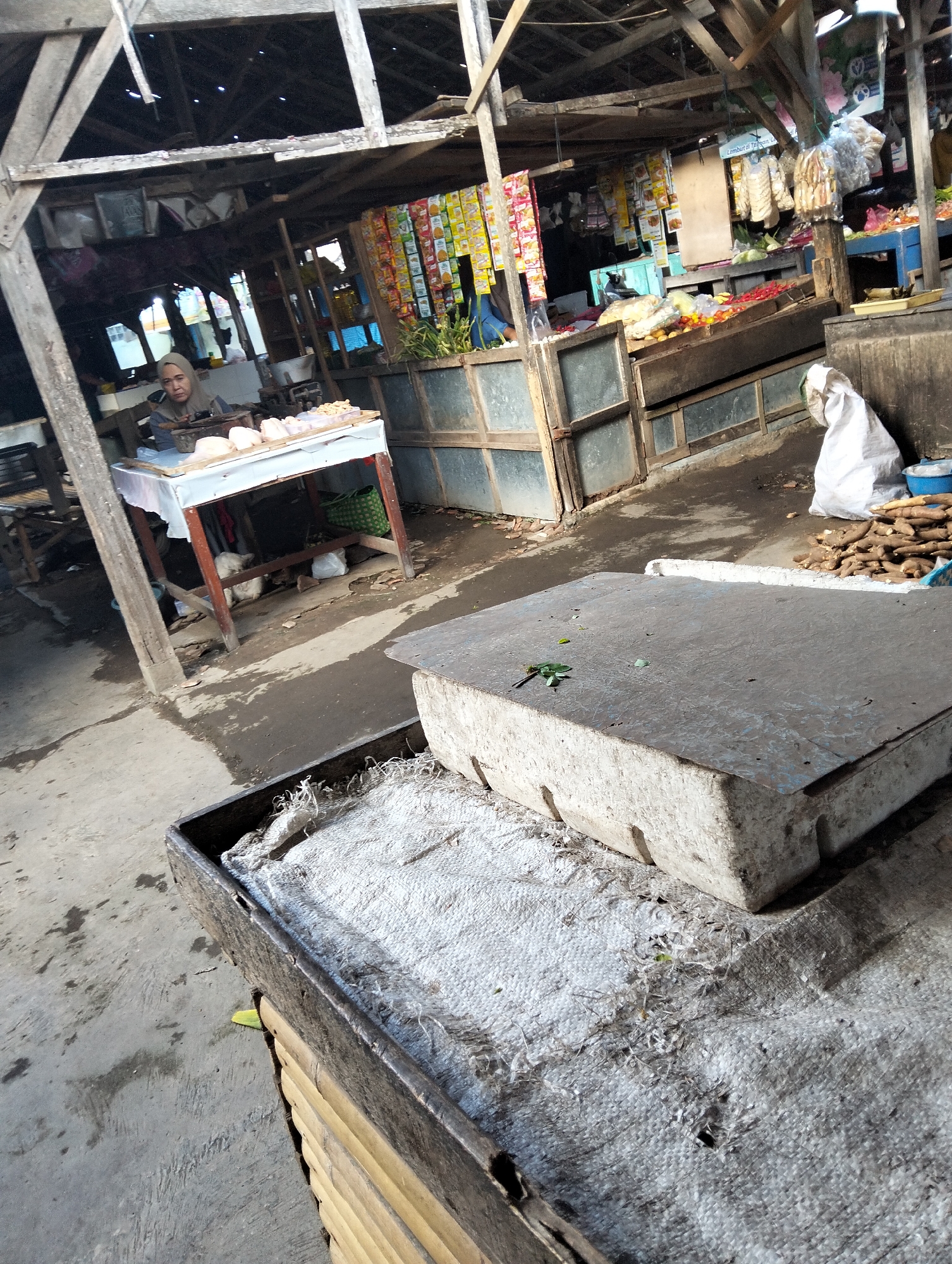 The image depicts a market stall in Muncar, Indonesia, likely focused on selling food items. In the background, a person wearing a hijab is seen behind a table laden with various food products, possibly dough or pastries. The stall has a rustic wooden structure with shelves displaying packaged goods and produce. In the foreground, a large concrete block rests on a textured surface, possibly a weighing scale or a display platform. The ground is paved with concrete and appears damp, with scattered debris. The overall scene suggests a bustling, open-air market environment typical of Indonesian towns.