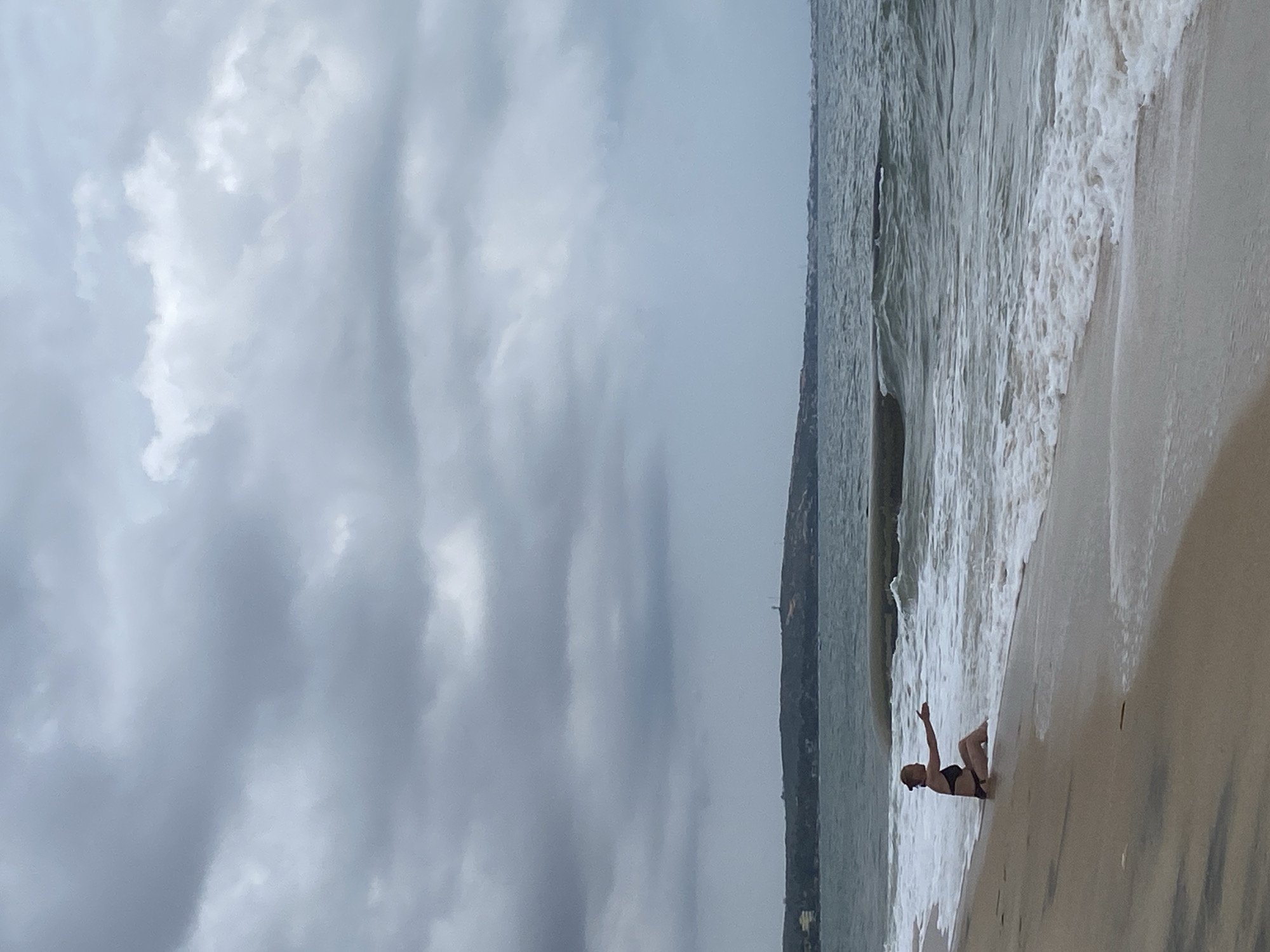 The image, originally rotated 90 degrees counter-clockwise, depicts a serene beach scene under an overcast sky. The sky, occupying a large portion of the frame, is filled with various shades of grey and white clouds, suggesting a cloudy day.
On the sandy shore, a single person, appearing to be a woman in a dark swimsuit or bikini, is lying or sitting at the water's edge. She is positioned with one arm raised and her legs bent, possibly relaxing or interacting with the incoming waves. The ocean water appears choppy, with white foam from breaking waves visible along the shoreline.
In the distance, across the water, a faint landmass or coastline can be seen, hinting at hills or low-lying structures. The overall lighting is soft and diffused due to the heavy cloud cover. The setting is a beach in Phu Thuy Ward, Vietnam. There is no visible text in the image.