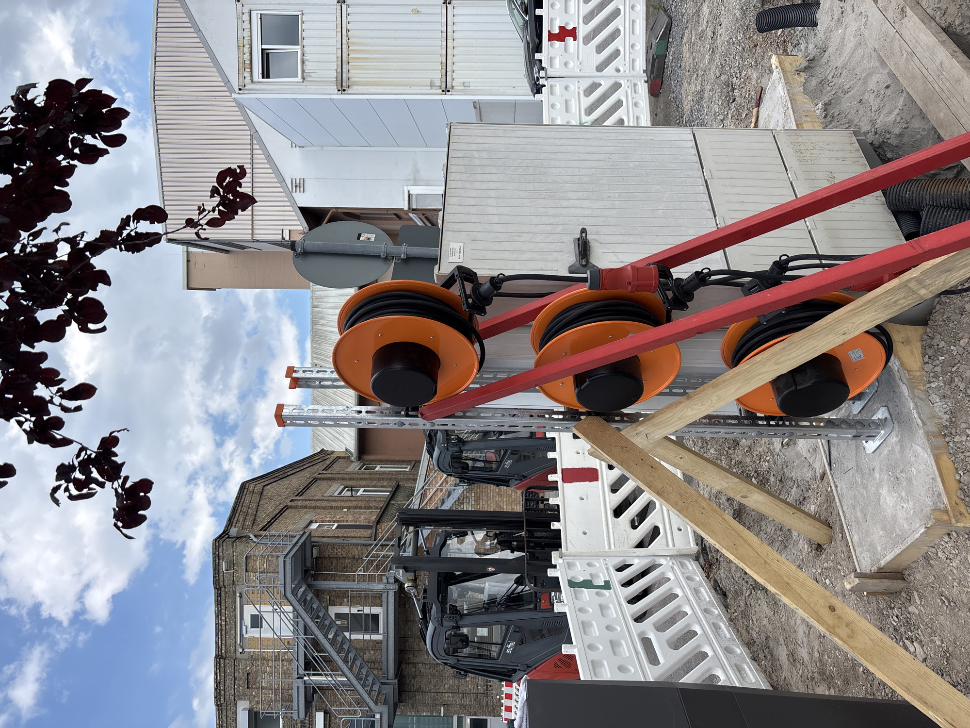 This media file shows a construction site in Mannheim, Germany. The scene is outdoors with a cloudy sky overhead. The perspective is from a slightly elevated angle, looking down and to the right.
Dominating the foreground are two large orange cable reels mounted on a red metal frame. The cables are black and appear to be heavy-duty electrical cords. These reels are suspended above a grey metal structure with multiple horizontal bars. Below this structure, two forklifts are visible. They are dark grey and red, with visible "RJHS" branding on their cabs.
In the background, there are several modular building units, likely temporary site offices or storage containers. These units are white and grey with corrugated metal siding and windows. A brick building with an external metal fire escape staircase is also visible to the left. A dark-leaved tree branch extends into the frame from the upper left, silhouetted against the sky.
The ground is a mix of gravel and concrete, with some wooden planks and white plastic barriers also present, suggesting ongoing construction work. The overall impression is one of active construction and utility work. There are no people visible in the image. The time of day appears to be daytime, given the brightness of the sky, though the clouds suggest it might not be a perfectly sunny day.