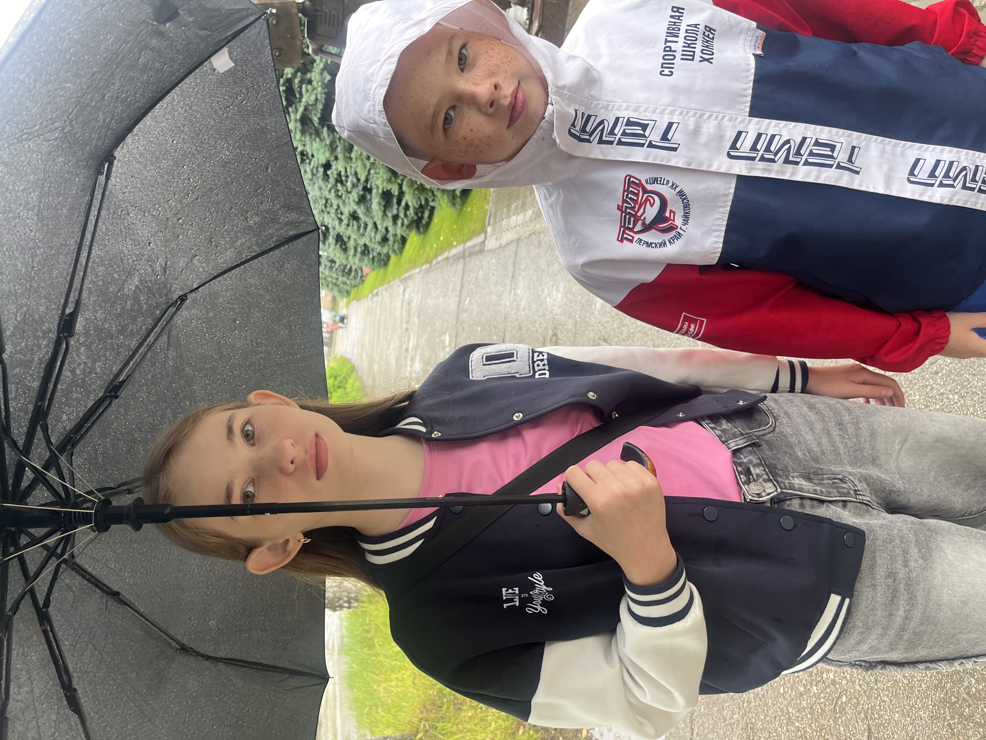 Two children, a teenage girl and a young boy, stand under a black umbrella on a paved path in an outdoor setting. The girl, on the left, has long brown hair and wears a pink t-shirt and a navy blue and white varsity jacket. The boy, on the right, has short blonde hair and freckles, and is wearing a white hooded jacket with red and blue accents. Both children appear to be looking towards the camera. The umbrella is open and has raindrops on its surface, indicating it is raining. The path is wet, and a row of evergreen trees lines the right side of the path. In the background, there are more trees and some indistinct figures walking. The setting suggests a public park or walkway in the city of Chaikovsky, Russia, given the visible text on the boy's jacket which reads "СПОРТИВНАЯ ШКОЛА ХОККЕЙ" (Sports School Hockey) and "ЧАЙКОВСКИЙ" (Chaikovsky).