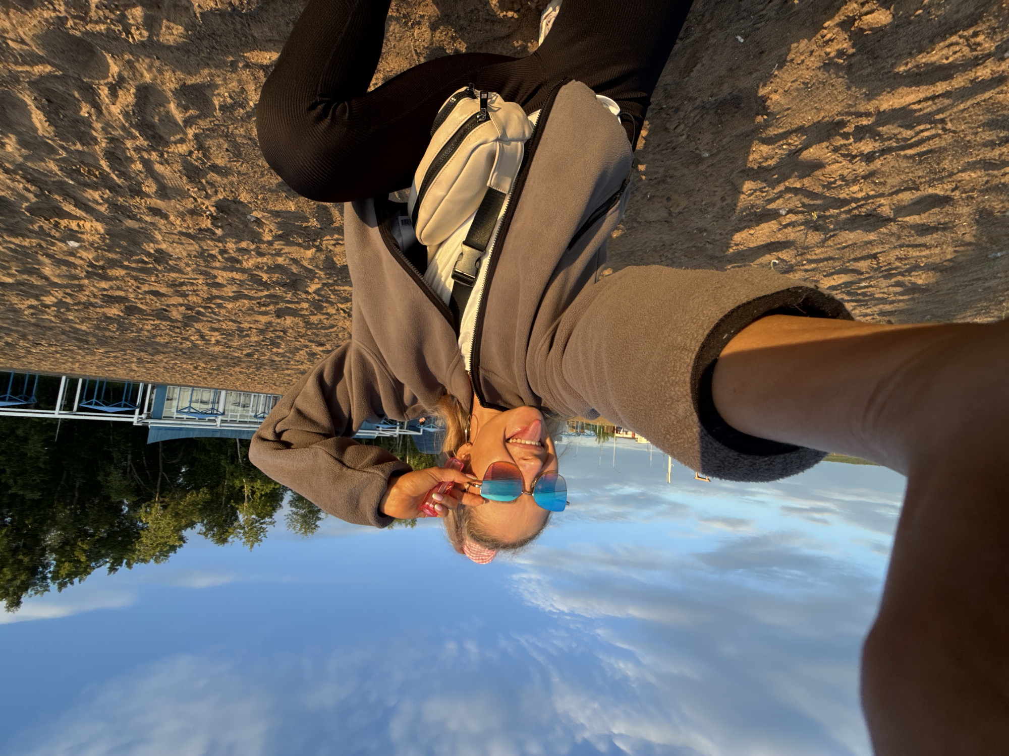 A young adult female is depicted in a wide-angle selfie, squatting on a sandy beach in Chaikovsky, Russia. She has blonde hair tied into a high ponytail with a pink and white checkered scrunchie and wears oversized blue and orange mirrored sunglasses. Her attire includes a grey zip-up fleece jacket, a white undershirt, and black ribbed pants. A beige fanny pack with black zippers and a black strap is worn across her torso. She extends her tongue and her left hand is positioned near her left temple, appearing to adjust her sunglasses. The scene is illuminated by golden hour sunlight, casting distinct, elongated shadows on the textured sand. In the background, light blue and white structures, possibly beach facilities, are visible, beyond which a dense line of green trees stands against a clear blue sky with scattered white clouds.