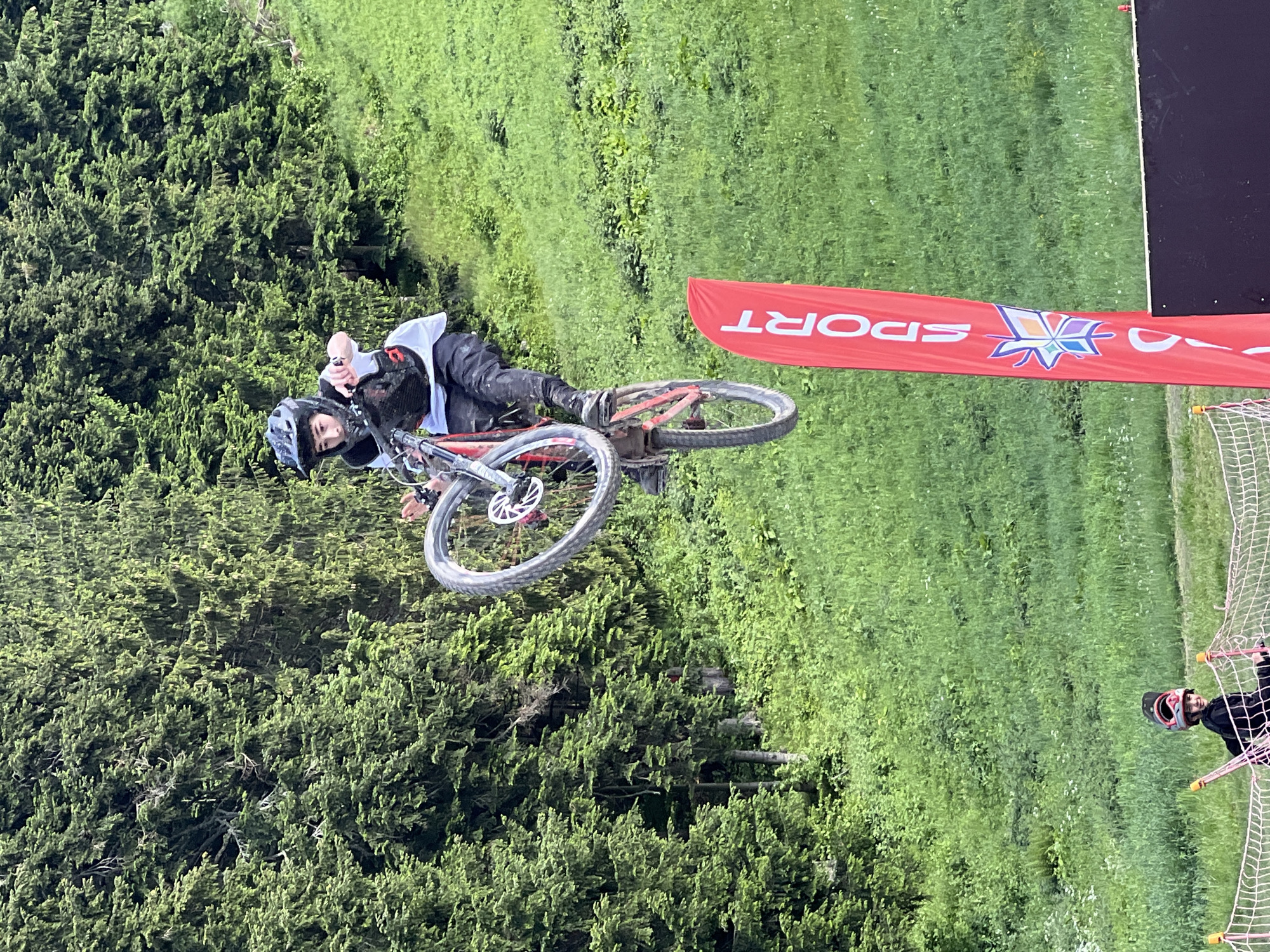 A person on a bicycle is airborne against a backdrop of green grass and trees. The person, wearing a helmet and protective gear, is on a red bicycle. The front wheel of the bike is angled upwards, with the rider's hands on the handlebars. A red flag with white lettering, including the word "SPORT," is visible in the foreground. In the lower left, another person is on the ground, also wearing a helmet. The scene appears to be an outdoor sports event or park.