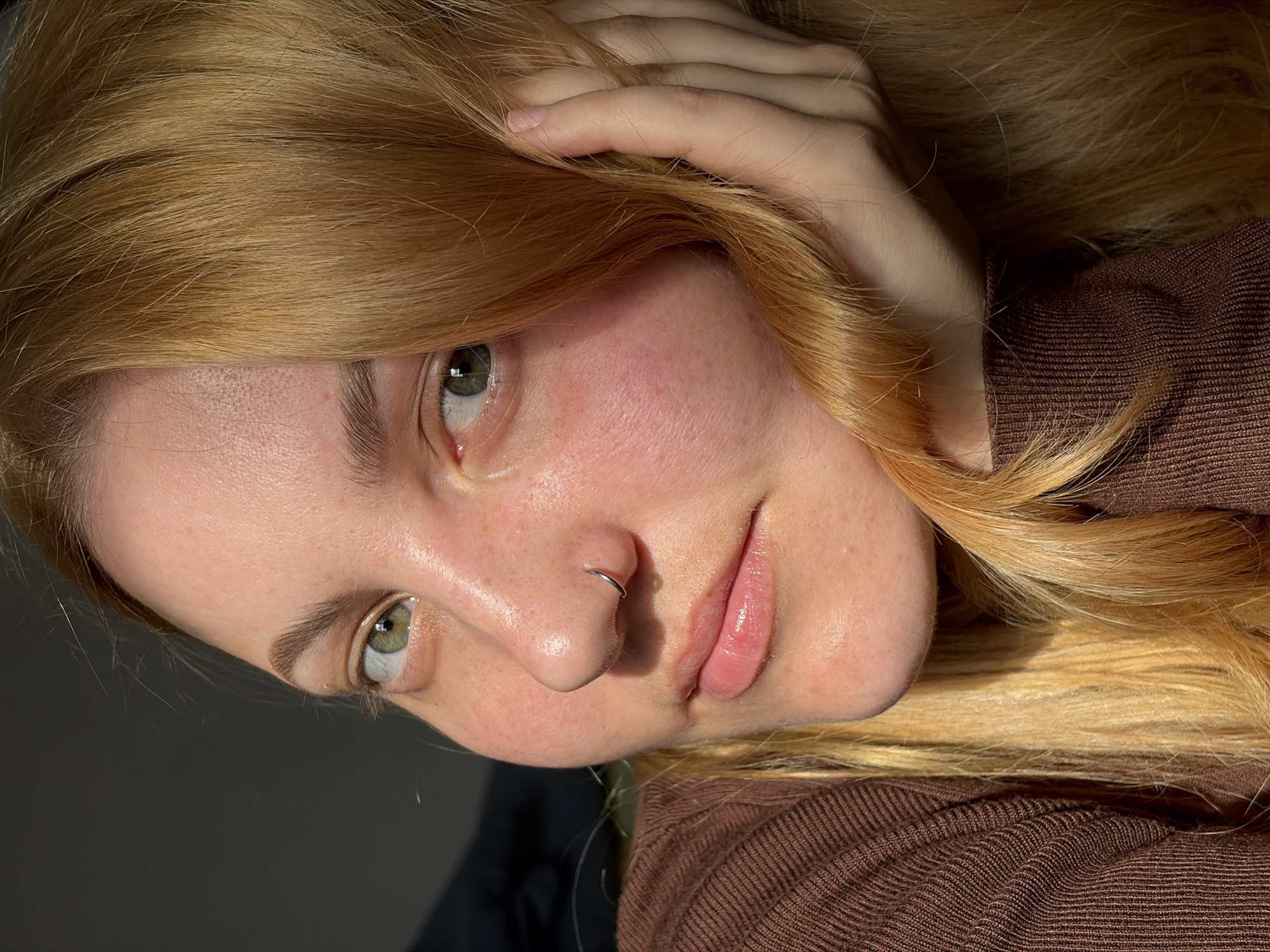 A young woman with light skin, green eyes, and blonde hair is pictured indoors. She has a nose ring. She is wearing a brown, ribbed-knit shirt or sweater. Her right hand is positioned near her head, likely touching her hair. The background is a mix of light and dark gray tones. The location is not apparent in this image and cannot be definitively identified as Izhevsk, Russia, or any other specific location.