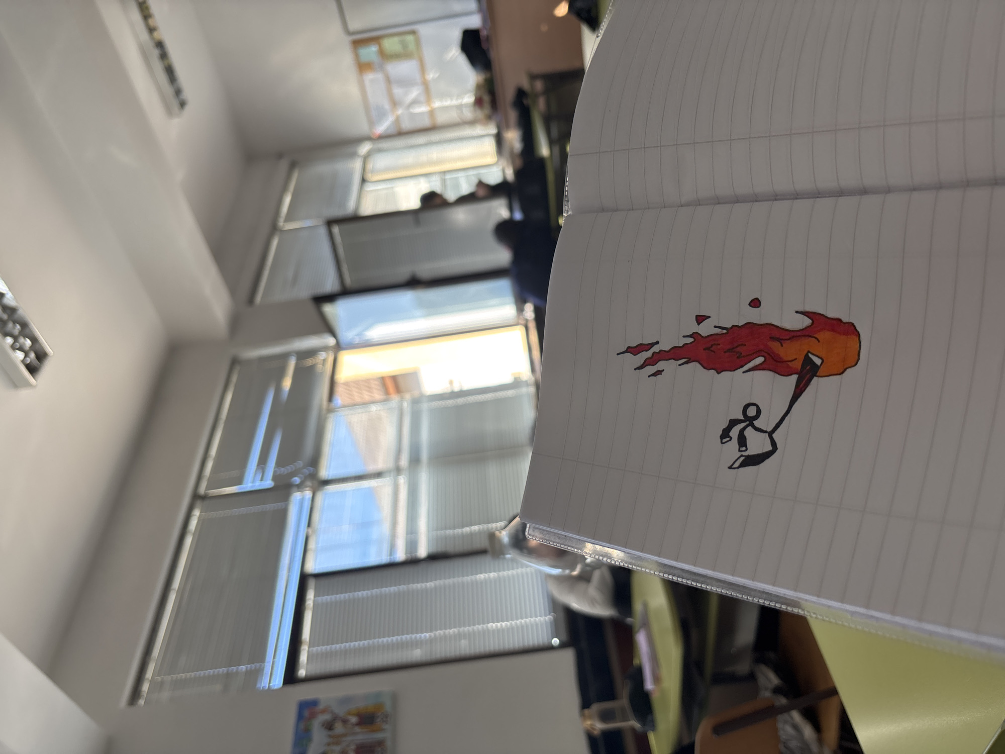 The image shows a notebook with a drawing of a stick figure holding a torch, viewed at an oblique angle. The background reveals a room with multiple windows covered with blinds, partially open, revealing a view of the sky. The room appears to be indoors, with a pale wall, a ceiling light, and what looks like a notice board on the far left. Other items and people are visible in the background, but are too blurry to identify. The image lacks any information to identify the location of the scene in the city of Zlatograd.