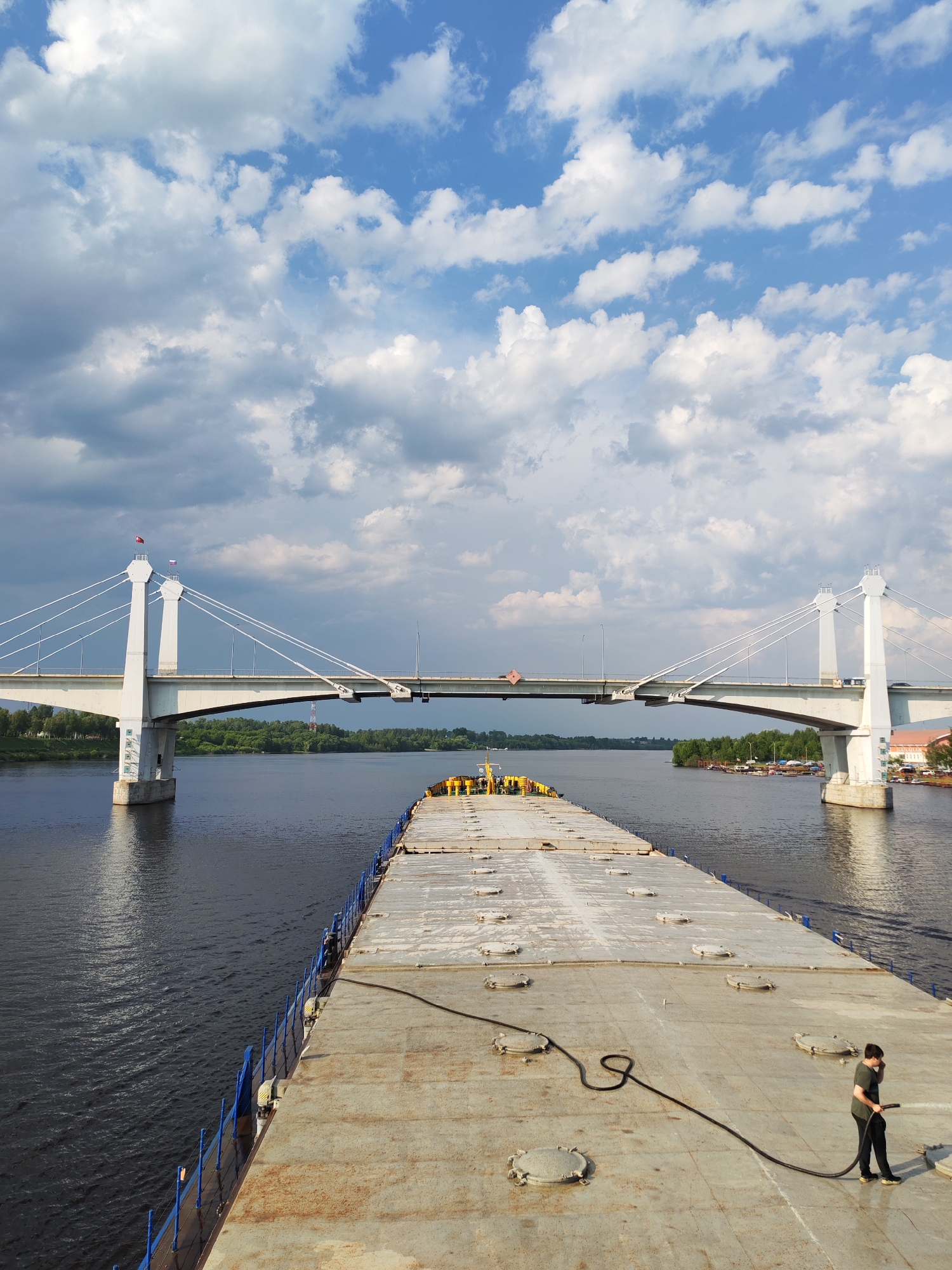 The image shows a long, flat-topped barge or pontoon boat traveling on a wide river, likely the Volga. In the foreground, a man is standing on the barge, holding a black hose. In the background, a cable-stayed bridge spans the river, supported by two white towers on either side. The bridge appears to be the Kimry Bridge in the city of Kimry, Russia, as it matches the known design and location. The sky is partly cloudy with patches of blue. There are trees visible on the riverbanks.