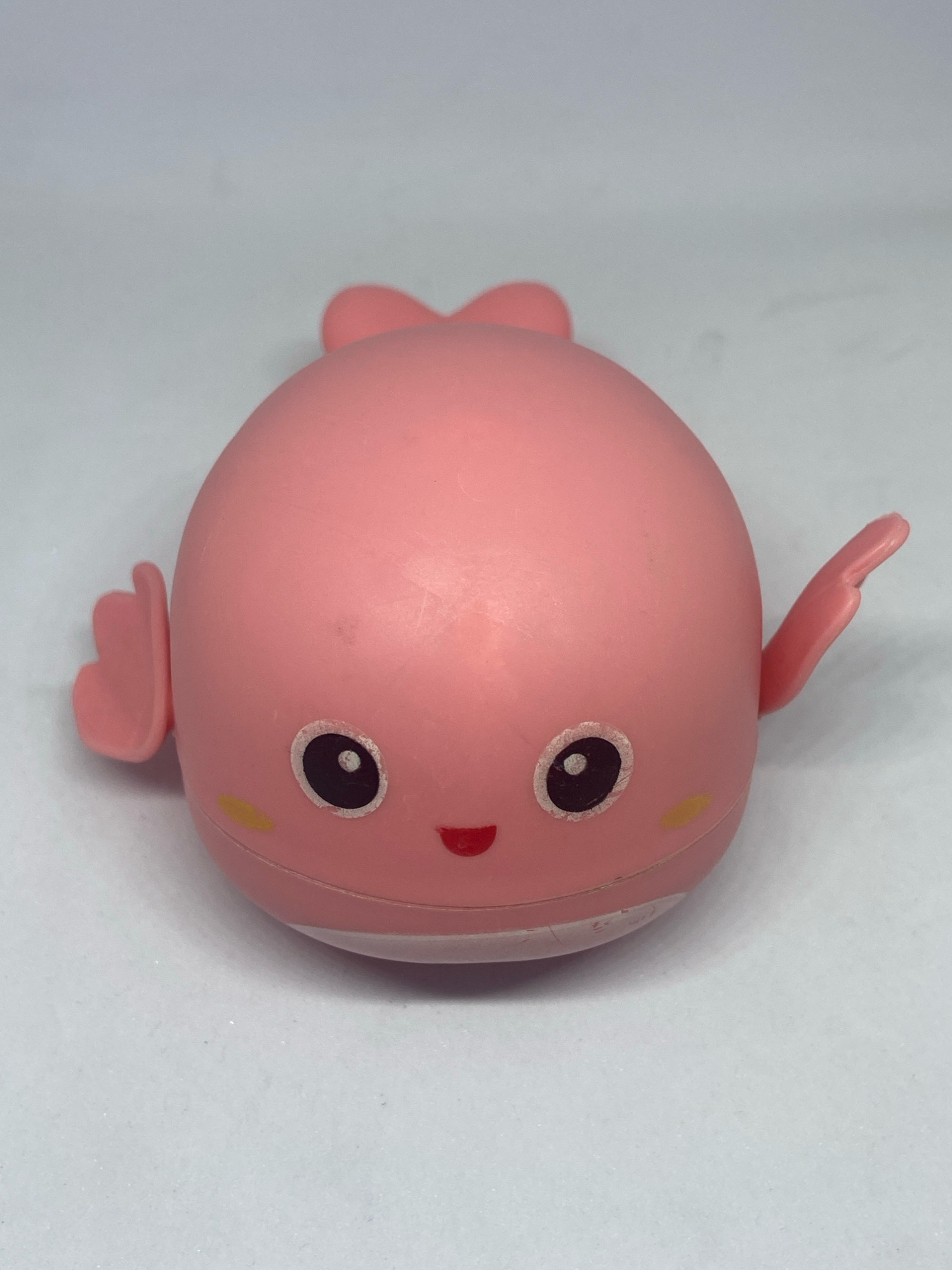 A pink toy whale is centered in the frame against a white background. The whale has a rounded body with a tail fin at the top. Two small pectoral fins extend from the sides of its body. The whale's face is adorned with black and white eyes, yellow cheek marks, and a smiling red mouth. There is a subtle white line around the bottom of its body. The image is a close-up, high-angle shot, suggesting it was taken in a studio or on a clean surface, not in any discernible outdoor environment within the city of Bekasi, Indonesia.