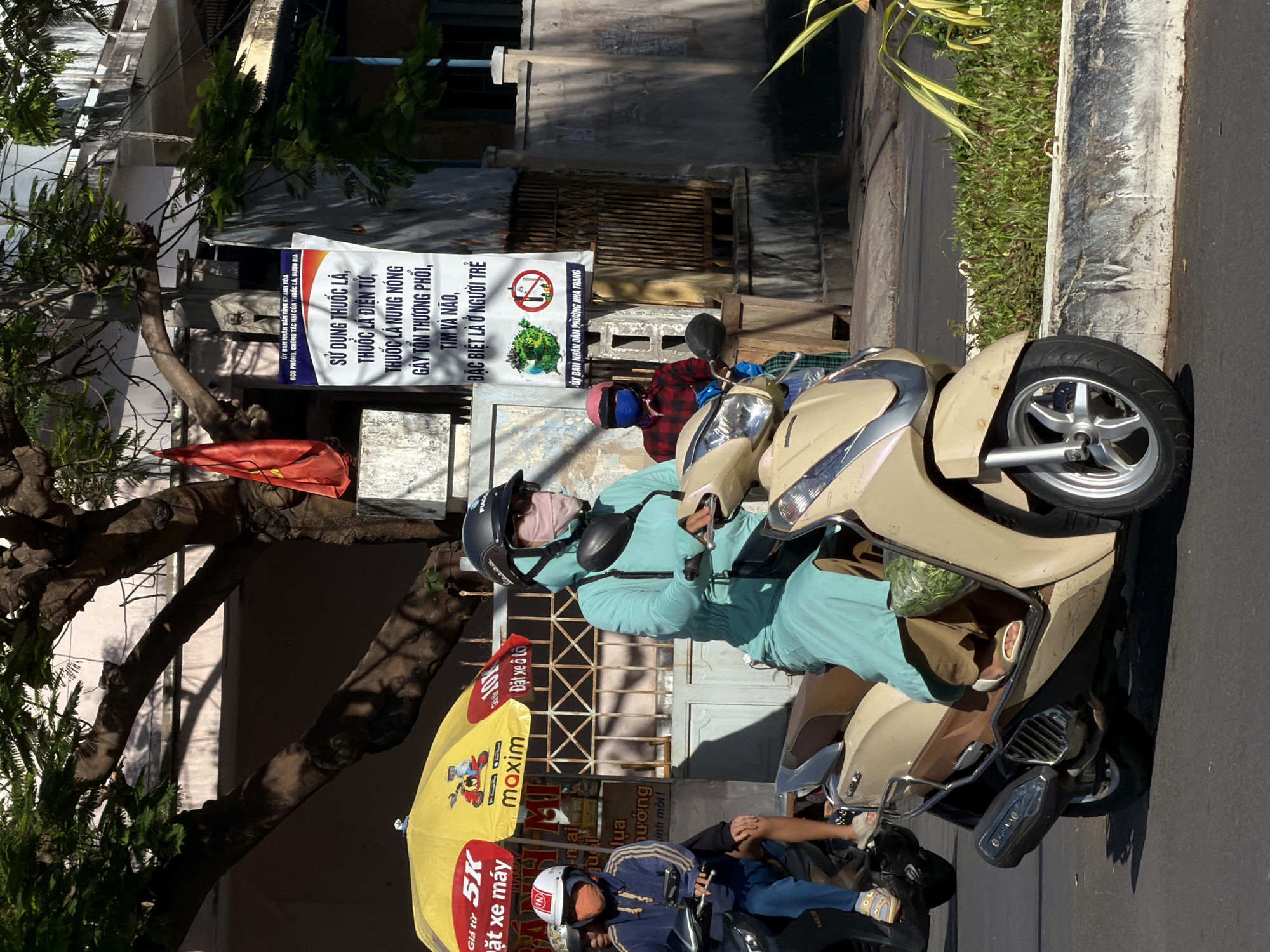 In Nha Trang, Vietnam, on a sunny day, two individuals are riding scooters. The foreground features a light-colored scooter with a rider wearing a turquoise long-sleeved jumpsuit, a face mask, and a helmet. They are carrying a bag of leafy green vegetables in the front compartment of the scooter. The rider's feet are visible, wearing sandals. Behind them, another scooter is partially visible with a rider wearing a blue jacket, white helmet, and an orange face mask. They are positioned behind a yellow umbrella with red text. In the background, a large tree with prominent branches shades a building with a pink facade. A red flag is visible hanging from a pole. A white banner with Vietnamese text and graphics, including an image of the Earth and a prohibition symbol over a syringe, is posted on a wall. The overall scene depicts a street in Nha Trang with typical urban elements like scooters, signage, and vegetation.