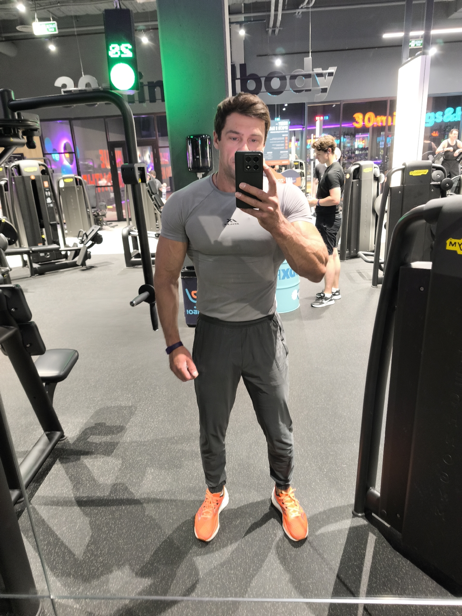 This media file shows a man taking a selfie in a gym. He is wearing a grey t-shirt and grey sweatpants, with bright orange running shoes. He has a watch on his left wrist and is holding a black smartphone in his right hand. The gym is well-lit with modern equipment, including weight machines and cardio equipment. In the background, other people are visible working out. There are also signs in the gym, one of which appears to read "30min" and another with the word "body." The overall atmosphere suggests a typical day at a fitness facility. The presence of artificial lighting and the indoor setting indicate that the time of day and weather are not discernible from the image. The location context provided is Moscow, Russia.