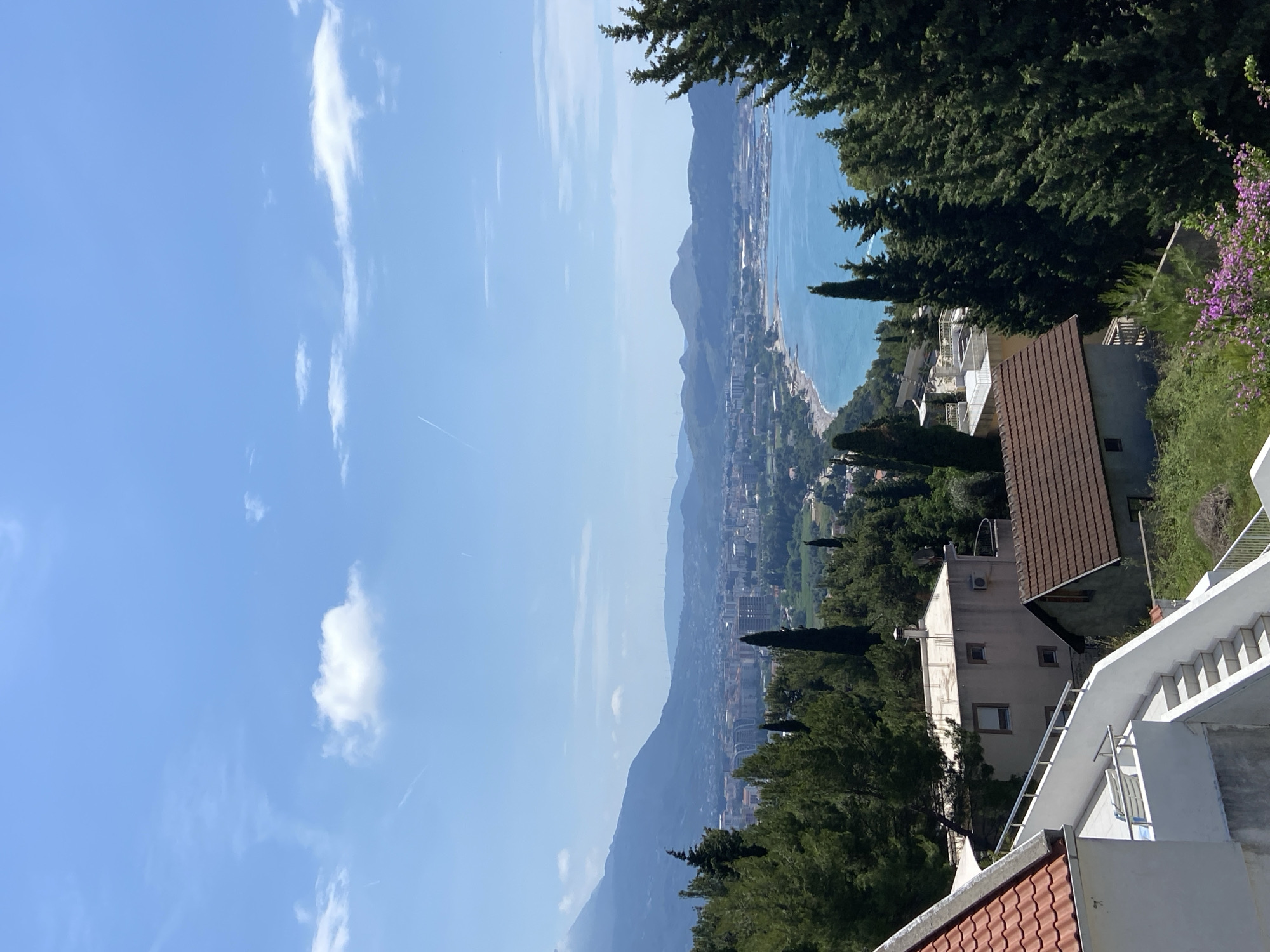 The image captures a view from Šušanj, Montenegro. The scene overlooks a coastline with the Adriatic Sea meeting a long beach and a cityscape that extends along the coast. Mountains rise in the background, creating a layered landscape. The sky is clear and blue with scattered white clouds. In the foreground, there are trees and parts of residential buildings, including tiled roofs and white walls. A staircase is visible on the right side of the frame, suggesting a tiered architectural structure.