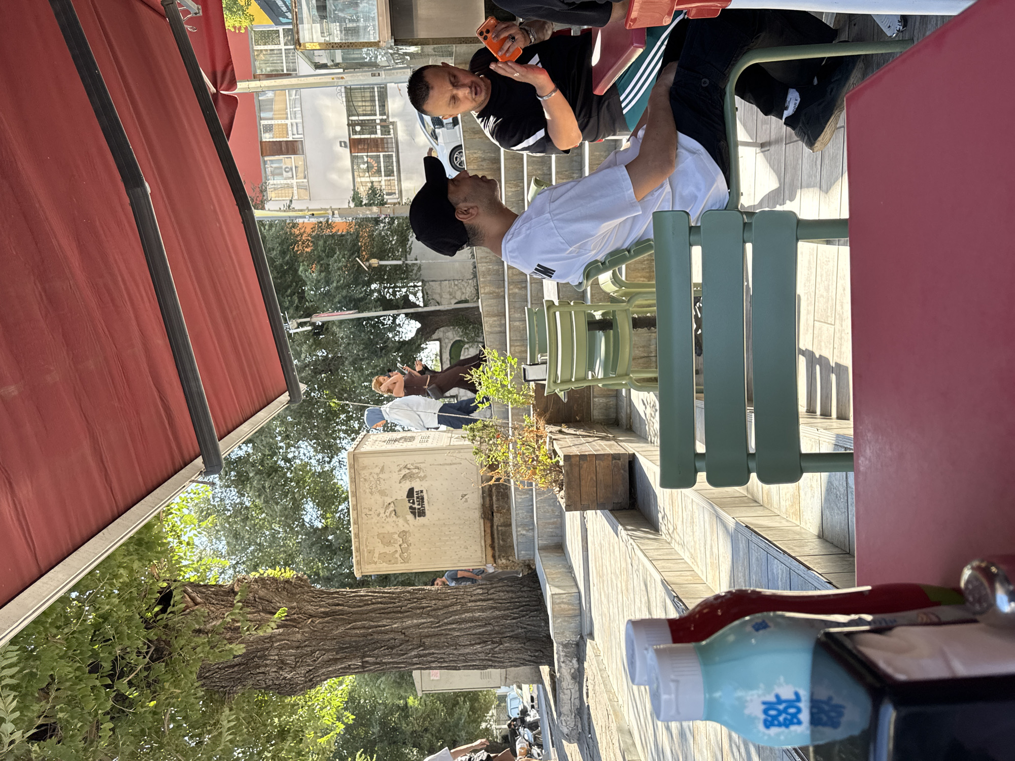 This is an outdoor scene at a cafe, likely in the Gungoren district of Istanbul, Turkey, given the context. Two men are seated at a table in the foreground. The man on the left wears a white t-shirt and a black baseball cap. The man on the right, wearing a black t-shirt, holds an orange mobile phone and appears to be gesturing with his right hand. They are seated on green metal chairs. A red table is visible in the lower foreground. In the background, there are steps leading up, a large tree with textured bark to the left, and several buildings. To the right of the steps, there are planters with some vegetation. Further back, a woman in a light blue headscarf and white top walks up the steps, followed by another woman in a brown outfit. The cafe is covered by a red awning. The ground appears to be paved.