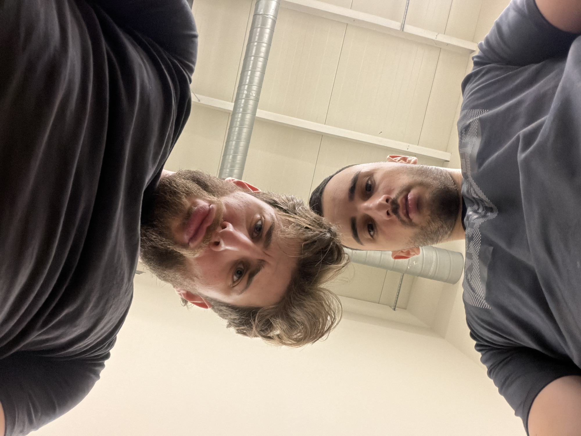 Two men are visible in an indoor setting under artificial lighting. The view is from above, looking down at the subjects. The man on the left has light brown hair and a beard, wearing a black shirt. The man on the right has short dark hair and stubble, wearing a gray shirt. The background consists of a white ceiling with metal pipes. It is impossible to determine if this image was taken in Plovdiv, Bulgaria without additional information.