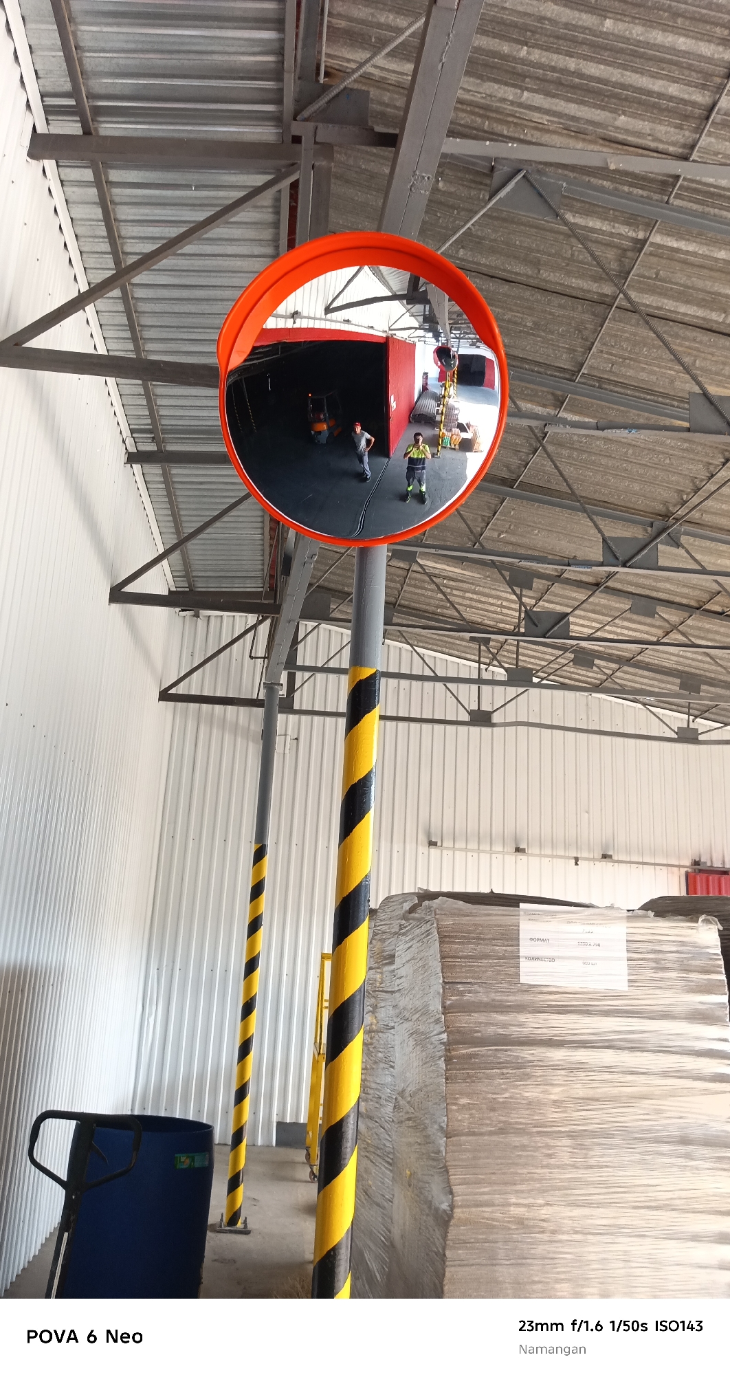 The image captures a wide-angle view inside a warehouse or industrial building. In the foreground, a large, orange-rimmed convex mirror is mounted on a gray pole striped with black and yellow bands. The mirror reflects the interior of the warehouse, showing two individuals wearing high-visibility clothing and a forklift. The environment appears to be a storage area, with large rolls of material wrapped in plastic visible on the right side of the frame. A blue industrial bin and a trolley are also present on the left. The ceiling of the building is made of corrugated metal, supported by a steel framework. The image is dated with camera settings: 23mm f/1.6 1/50s ISO143, and the location is identified as Namangan.