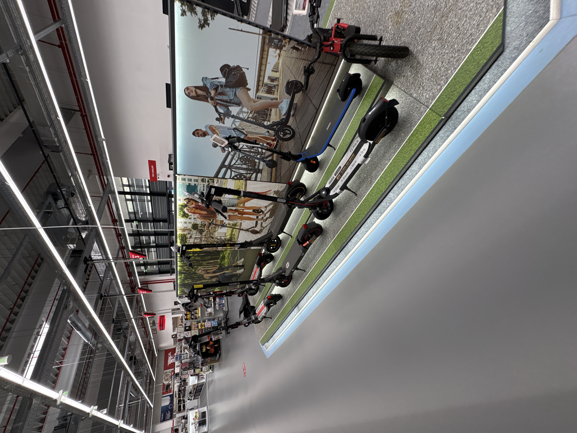 The image depicts the interior of a store, potentially a department store or electronics retailer, in what appears to be the city of Lovech, Bulgaria. A series of electric scooters are prominently displayed on a platform designed to resemble a sidewalk or grass strip. Behind the scooters, a large advertisement panel showcases images of people riding similar scooters in urban settings. The scooters themselves vary in design and color, featuring brands like Segway and Xiaomi. Above, the store's ceiling is visible, with industrial lighting fixtures and metal support structures. Aisles of shelves stocked with various products are visible in the distance, suggesting a wide variety of goods are available. The overall impression is of a well-lit retail environment offering personal transportation devices.