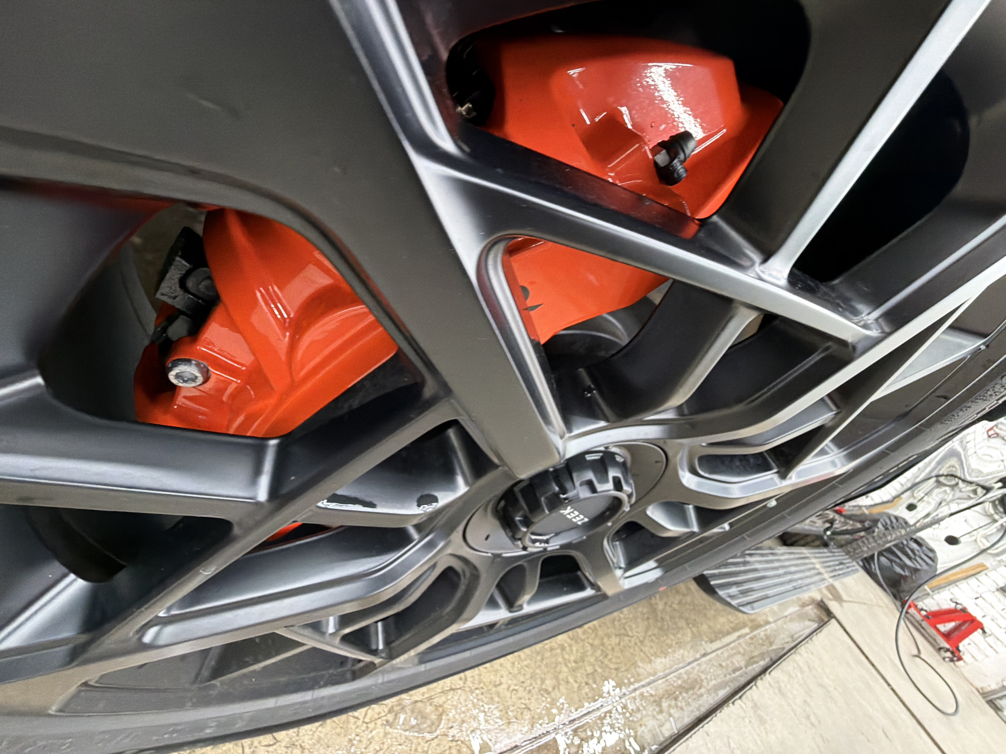 The image shows a close-up of a car wheel with orange brake calipers. The wheel is a dark gray color with an intricate, multi-spoke design. A black center cap with the word "ZEEK" is visible. The vehicle is on a lift in what appears to be a garage or auto repair shop. The floor is wet. A red jack and other equipment are partially visible in the background. The location cannot be determined from the image.