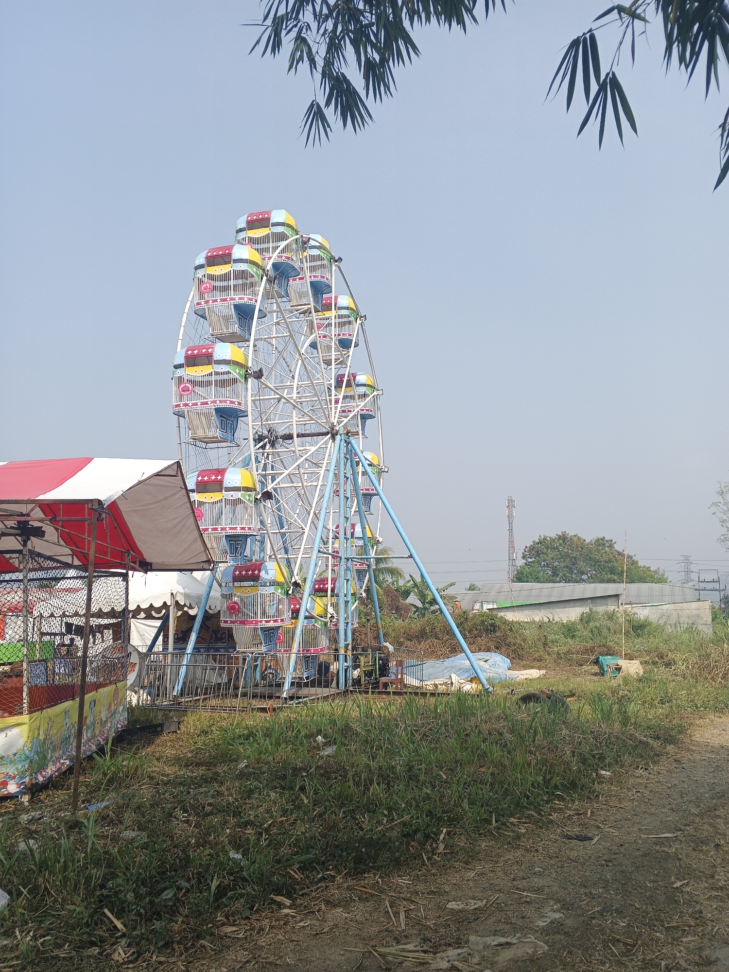 In the city of Sukabumi, Indonesia, a moderately sized Ferris wheel with colorful passenger cabins stands prominently on a dirt path. The Ferris wheel, constructed of white metal with blue supports, is partially obscured by the lush green grass and dry vegetation surrounding it. To the left of the Ferris wheel, a striped red and white tent is set up, accompanied by a fenced-off area that appears to be a vendor stall. In the background, buildings and utility poles are visible, indicating a semi-urban or transitional environment. The sky is clear and pale blue, with the branches of a tree visible in the upper right corner of the frame.