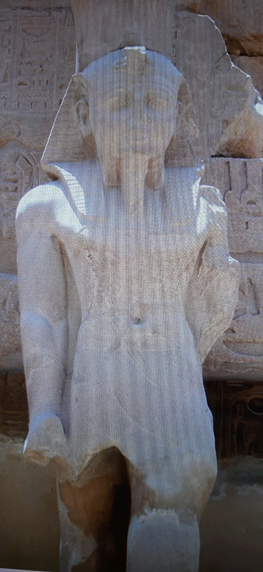 The image shows a close-up of a weathered, light-colored stone statue of an ancient Egyptian pharaoh. The statue is partially illuminated, with light falling on its upper body and head. The pharaoh wears a nemes headdress and a false beard, characteristic of royal iconography. The statue is in front of a stone wall covered in hieroglyphs, which are partially visible. The stone appears to be sandstone or limestone. The statue's surface is eroded, with some areas showing discoloration and pitting. There is no visible context to suggest this statue is located in Chernihiv, Ukraine, and it is highly unlikely given the subject matter. The statue appears to be standing, but only the upper torso and head are visible in the frame. The overall impression is one of antiquity and historical significance.