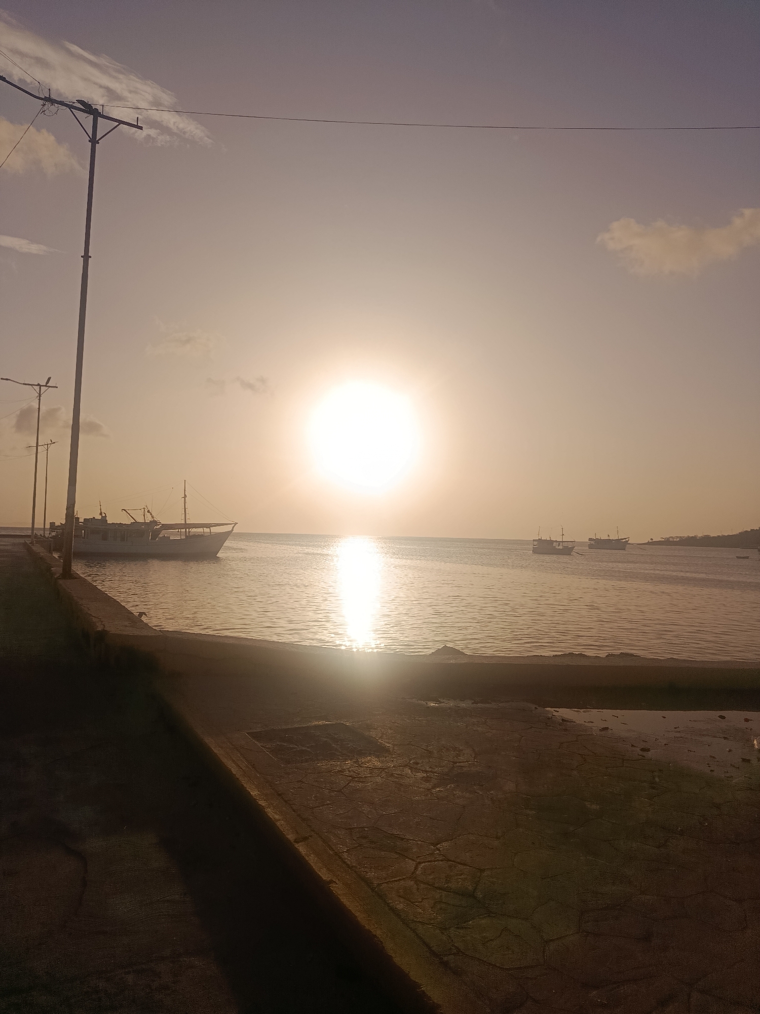 The image shows a waterfront scene at sunset in Juan Griego, Venezuela. The sun is low on the horizon, casting a warm glow over the water and sky. Several boats are anchored in the bay, and a pier or promenade lines the foreground. The water is calm, reflecting the light of the setting sun. The sky is a soft gradient of pink and orange, with a few wispy clouds. The scene appears serene and peaceful, capturing the beauty of a coastal sunset.
