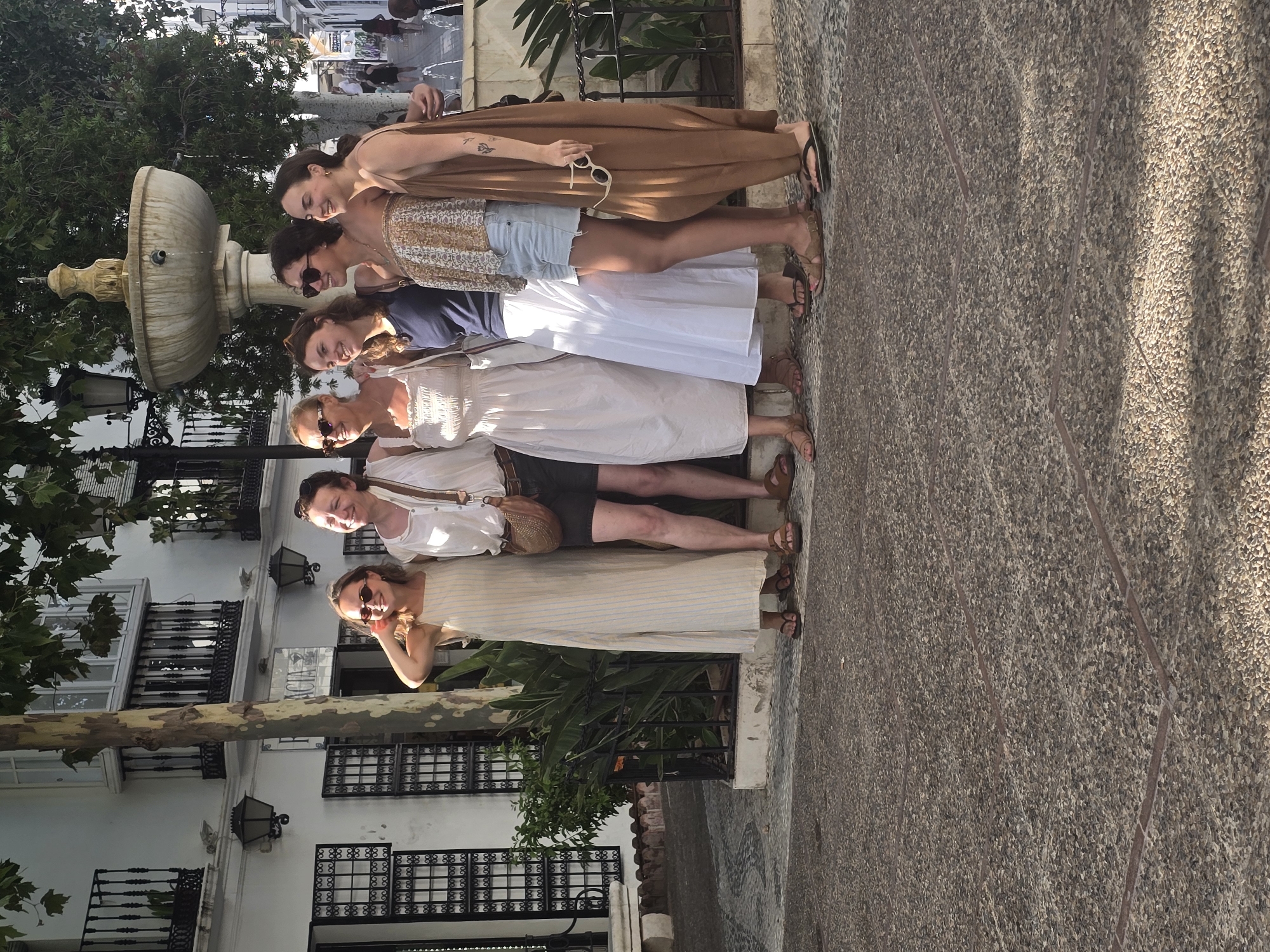 A group of six young women are posing for a photograph in a European town square. The woman on the far left is wearing a cream-colored dress with thin blue stripes, and she has blonde hair and sunglasses. To her right, a woman in a white sundress smiles at the camera. The third woman from the left wears a blue tank top and denim shorts, her hair pulled back. The fourth woman from the left is wearing a strapless patterned top, denim shorts, and sunglasses. The fifth woman from the left is wearing a brown jumpsuit and holding a pair of white sunglasses. The woman on the far right is wearing a brown dress. All six women are wearing sandals. Behind them, a stone fountain with a tiered design is visible. The ground is paved with small stones. The buildings in the background are whitewashed with wrought-iron balconies. A tree with rough bark stands to the left of the fountain. The scene appears to be in Mijas Pueblo, a white village in Andalusia, Spain.