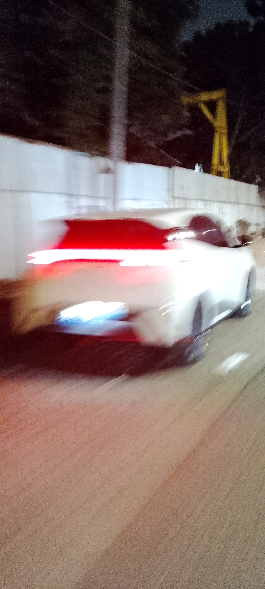 The image, taken at night in Bekasi, Indonesia, depicts a motion-blurred white car with its taillights illuminated in bright red. The car is driving on a road, possibly a highway, indicated by the white dashed lines visible on the asphalt. In the background, a white wall runs horizontally along the side of the road, and beyond that, dark foliage can be seen. A tall, thin utility pole stands to the left of the frame, partially obscuring the sky, which is dark. To the right, a yellow, crane-like structure is partially visible against the night sky. The overall impression is of a vehicle in motion at night in an urban or semi-urban environment.