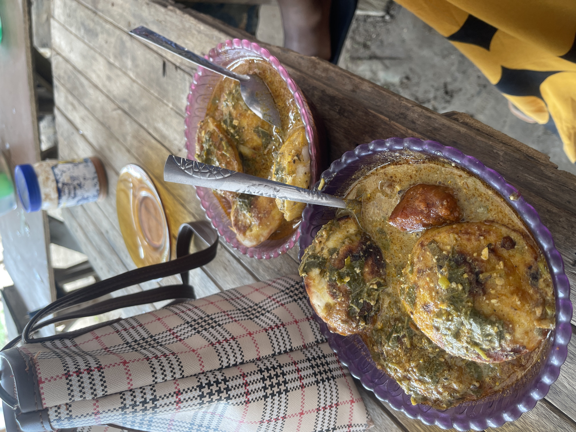 Two purple and pink plastic bowls of food, likely some sort of stew with pieces of boiled eggs, sit on a rough wooden table outdoors. Spoons are in each bowl. To the left of the bowls is a patterned tote bag with brown straps. In the background, there is a partially visible yellow plate and a blue-capped bottle. The scene is likely a casual outdoor dining setting in Rafin Par, Nigeria.