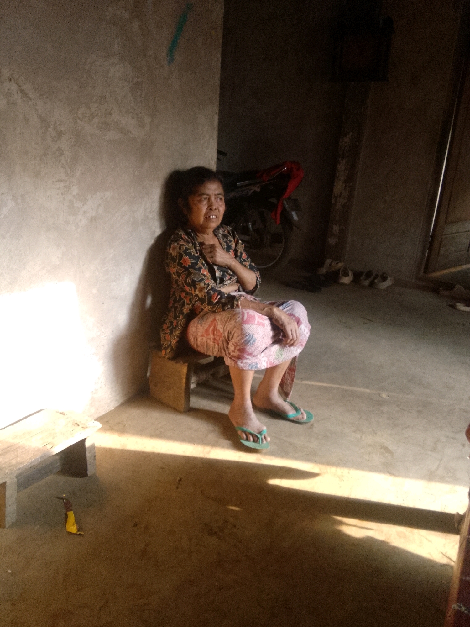 A middle-aged Indonesian woman sits on a wooden stool in an unlit room with a concrete wall on the left. She is wearing a floral patterned shirt and patterned pants. Her feet are clad in flip-flops. Sunlight streams in from the left, casting a strong shadow across the floor. In the background, a red object, possibly part of a motorcycle, is visible. Several pairs of shoes are scattered on the floor in the background. The overall environment appears to be humble and sparsely furnished.