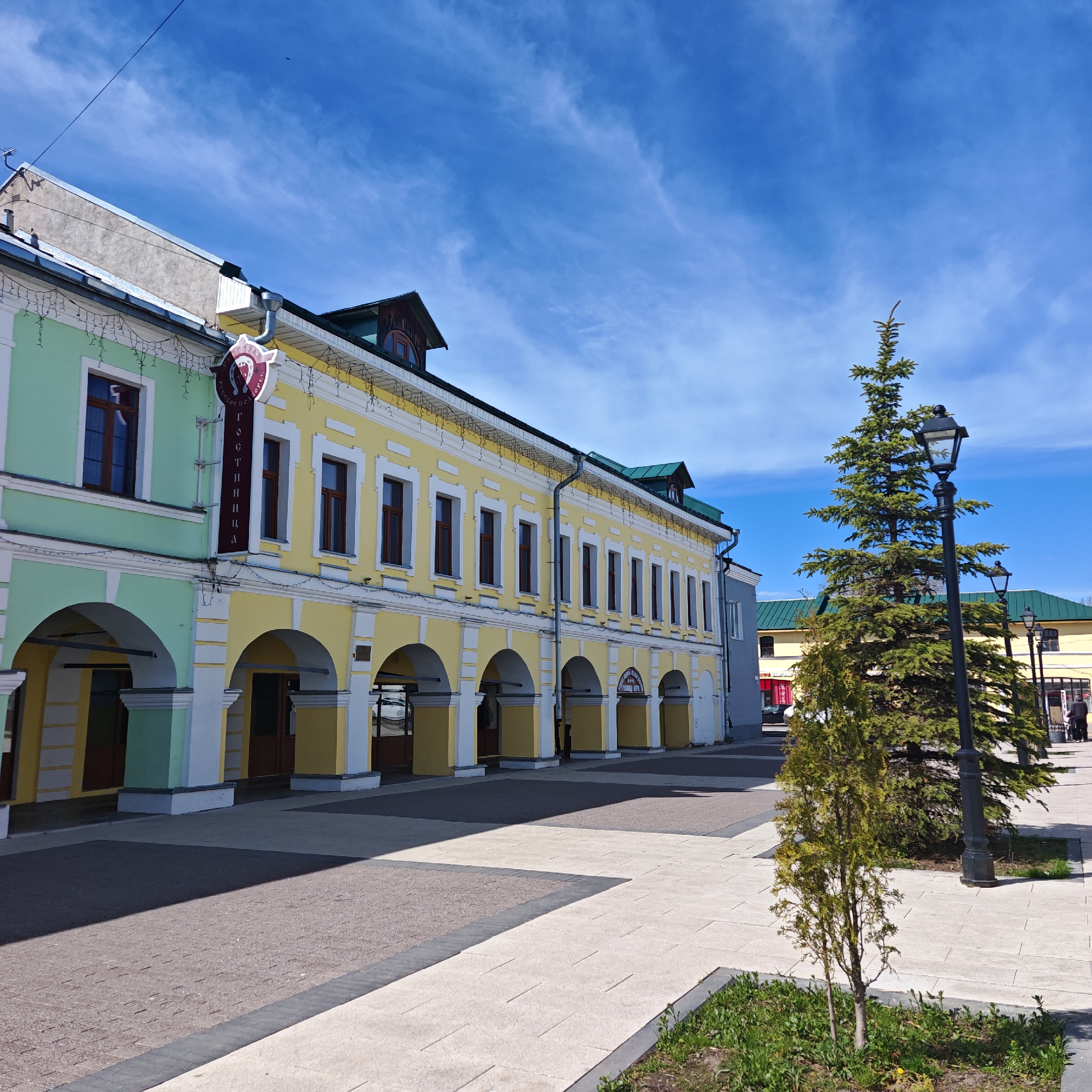 The image depicts a street scene in Rostov, Russia, showcasing a two-story building with a green and yellow facade, characterized by arched walkways supported by columns. A sign reading "гостиница" (hotel) is vertically displayed. A cobblestone and light-colored tiled pedestrian area extends towards the foreground. A young evergreen tree and a black ornate streetlight are positioned on the right side. In the background, other buildings with green roofs can be seen. The sky is blue with scattered clouds. Some people are visible in the background near the building with the green roof.
