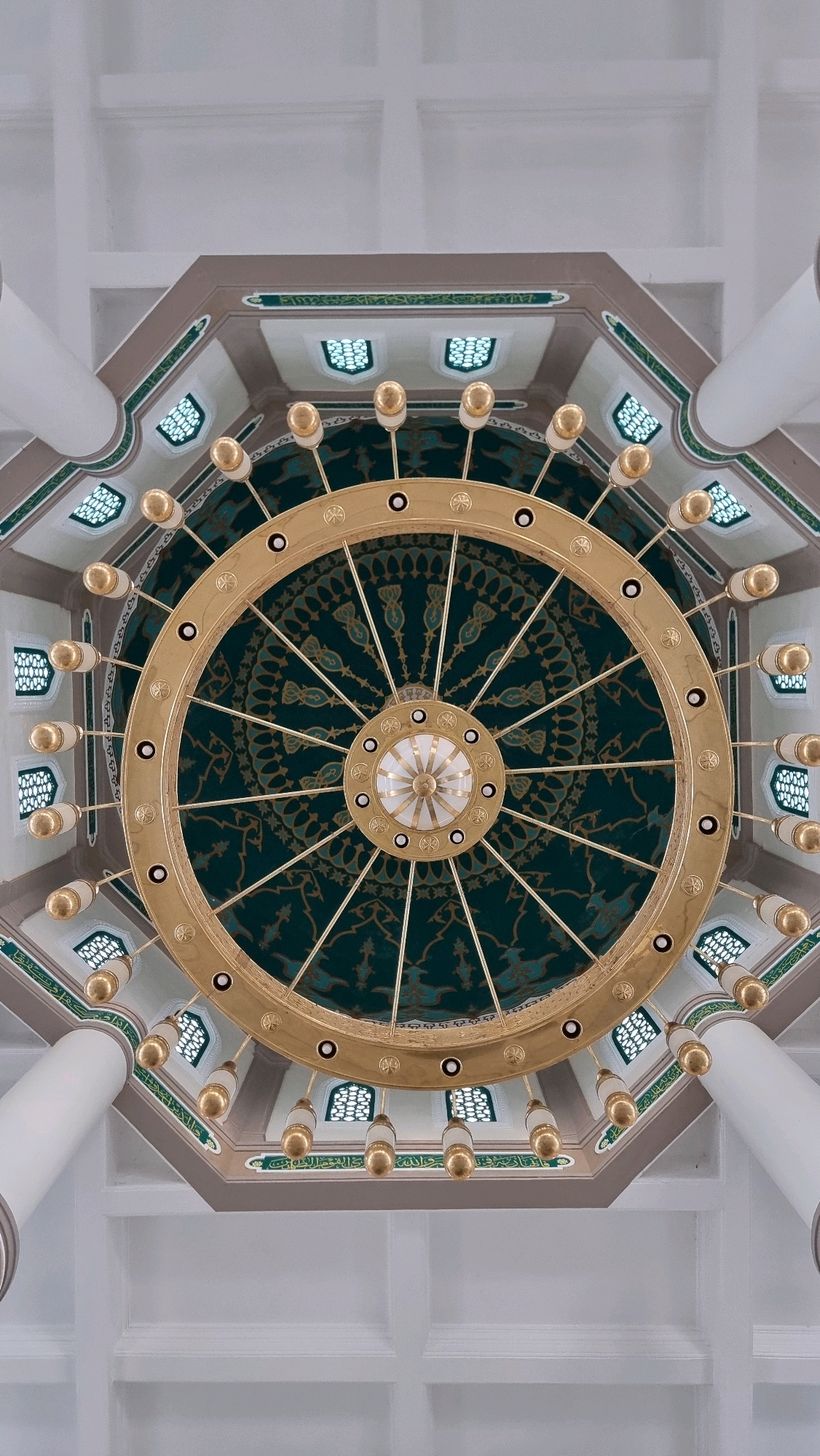 The image shows a low-angle, centered shot of a large, ornate chandelier hanging from the ceiling of a building, likely a mosque or Islamic cultural center. The chandelier is circular and made of gold-colored metal, with numerous bulbs or light fixtures extending outwards. The ceiling itself is octagonal, with decorative green and gold patterns and Islamic calligraphy. Several white columns are visible, supporting the ceiling. The overall impression is one of grandeur and intricate craftsmanship. This scene is located within the Baiturrahman Grand Mosque in the city of Lhokseumawe, Indonesia.