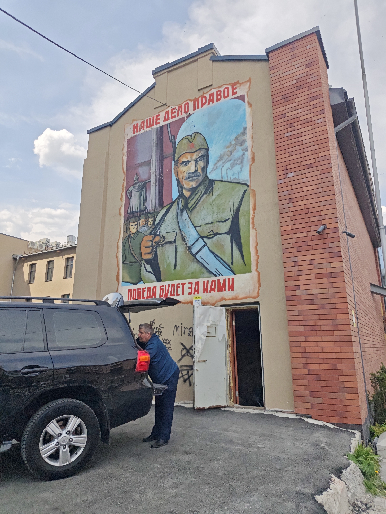 A man is looking into the open trunk of a black SUV, which is parked in front of a building. The building has a large mural of a Soviet soldier on its facade, which also has text in Russian that translates to "Our cause is just, victory will be ours." The mural appears to be painted on a beige wall, which is adjacent to a brick wall. The man is wearing a blue shirt and dark pants. The building has an open door next to the mural. The location is Yekaterinburg, Russia.