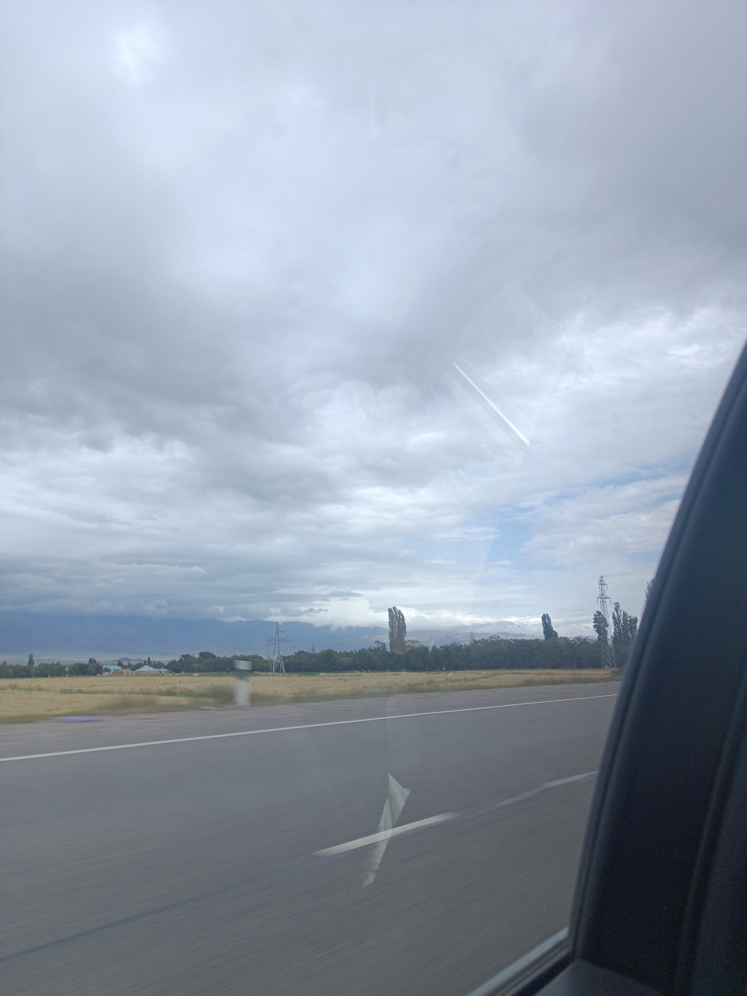 The image captures a view from a vehicle moving on a paved road, likely in the vicinity of Sasyk-Bulak, Kyrgyzstan, given the provided context. The foreground is dominated by the asphalt road, marked with white lines, indicating a direction of travel. The right side of the frame shows a portion of the vehicle's interior, possibly a window frame and part of a car door, suggesting the perspective is from inside a moving car.
Beyond the road, a flat, grassy landscape stretches out. In the mid-ground, there are scattered trees and a few low-rise buildings, suggesting a semi-rural or suburban environment. A prominent feature in the distance is a high-voltage power line tower. Further in the background, a range of hills or mountains can be observed under a heavy, overcast sky. The sky itself is filled with a mix of gray clouds, with some patches of lighter sky visible, hinting at diffused daylight. Two faint, diagonal white streaks in the sky suggest contrails from an aircraft. The overall atmosphere is subdued due to the cloud cover. There are no people visible in the image.