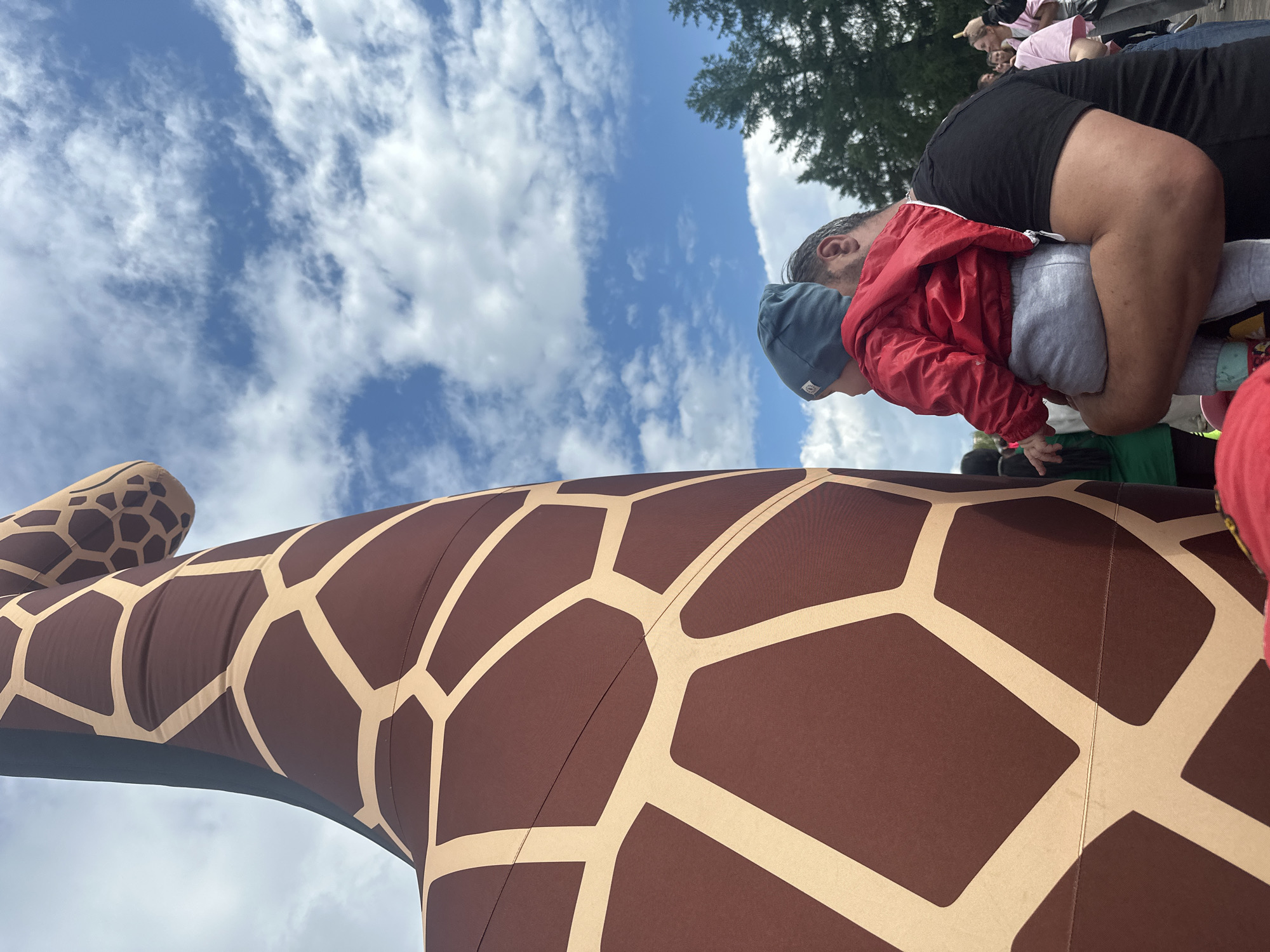An outdoor medium shot captures a man holding a baby in his arms, with the side of a giant inflatable giraffe dominating the left half of the frame. The giraffe is brown with a light tan pattern of irregular polygons. The baby, facing away from the camera, wears a blue beanie and a red jacket over gray pants. The man, also facing away, has dark hair and is wearing a black t-shirt. Behind them, a line of trees is visible against a blue sky dotted with white clouds. Other people are faintly visible in the background, suggesting a public gathering or event. The scene appears to be in an open area, possibly a park or festival ground, in an unspecified urban location.