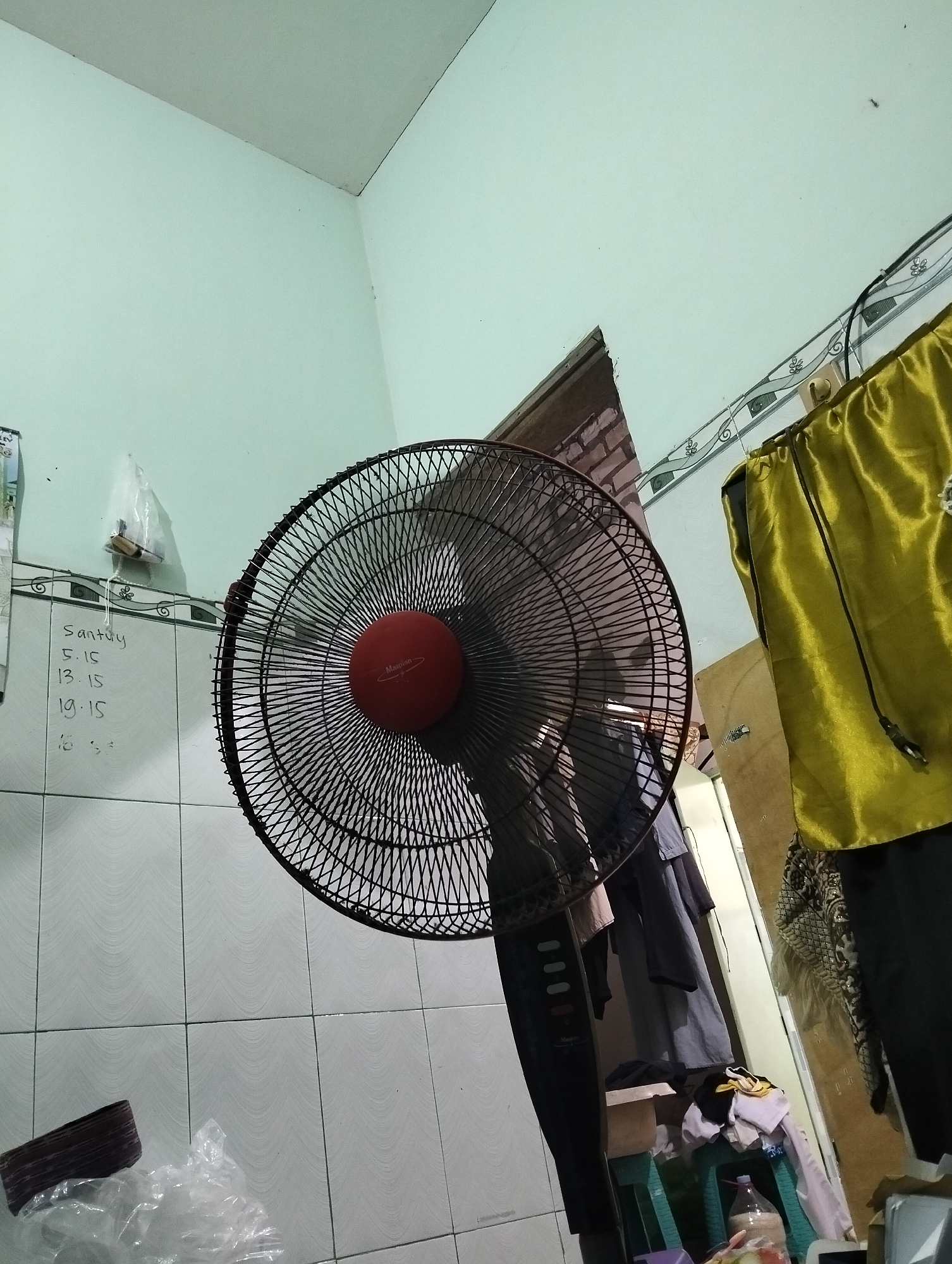 This image depicts an interior scene, likely a bedroom or living area, in Paciran, Indonesia. The focal point is a black oscillating fan with a red center, positioned slightly off-center to the right. To the left of the fan, on a tiled wall, a list is written in what appears to be marker, containing the words "Santuy" and several time stamps: "5.15", "13.15", "19.15", and partially obscured numbers. A small plastic-wrapped item hangs on a string above the list.
The background reveals a pale green wall meeting a white ceiling. To the right of the fan, a bright yellow fabric, possibly clothing or a curtain, is draped. Further to the right, a doorway or opening shows a stack of clothes on a green stool, with a plastic bottle partially visible. The floor is tiled with a light pattern. The overall impression is one of a lived-in space with a functional layout.