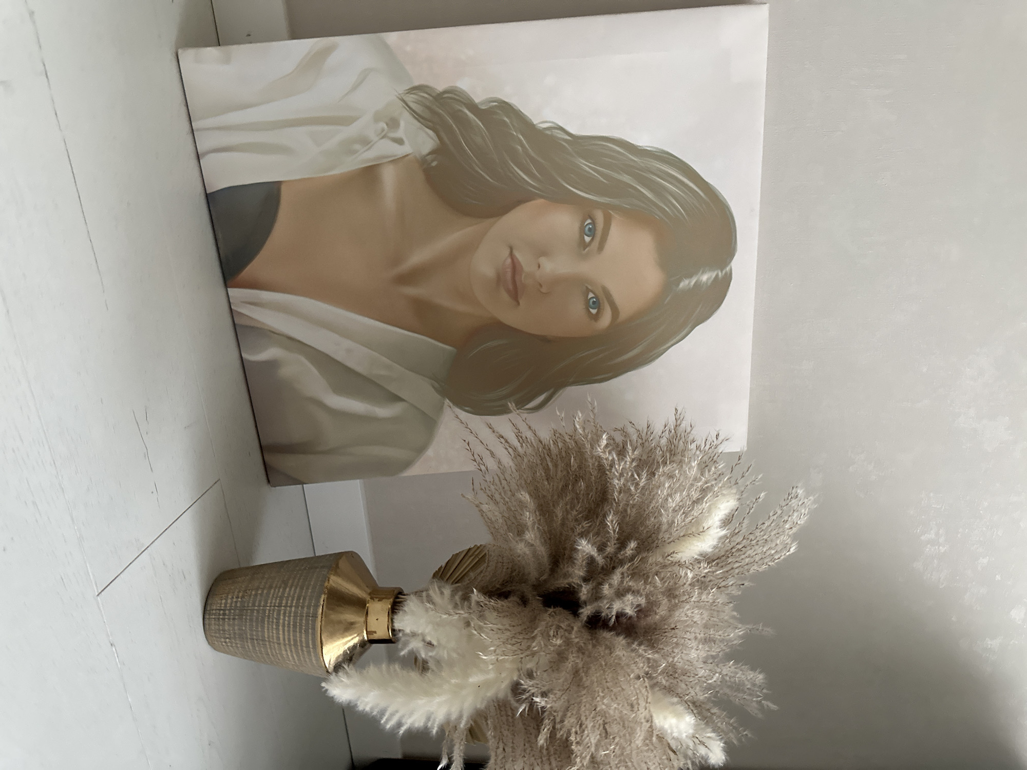 A framed portrait of a woman with dark hair and blue eyes wearing a white shirt is propped against a white wall with wood-look tile flooring. In front of the painting is a brown and gold vase filled with dried grasses. The location is stated as Прикамский, Russia, though there are no contextual clues to support this.