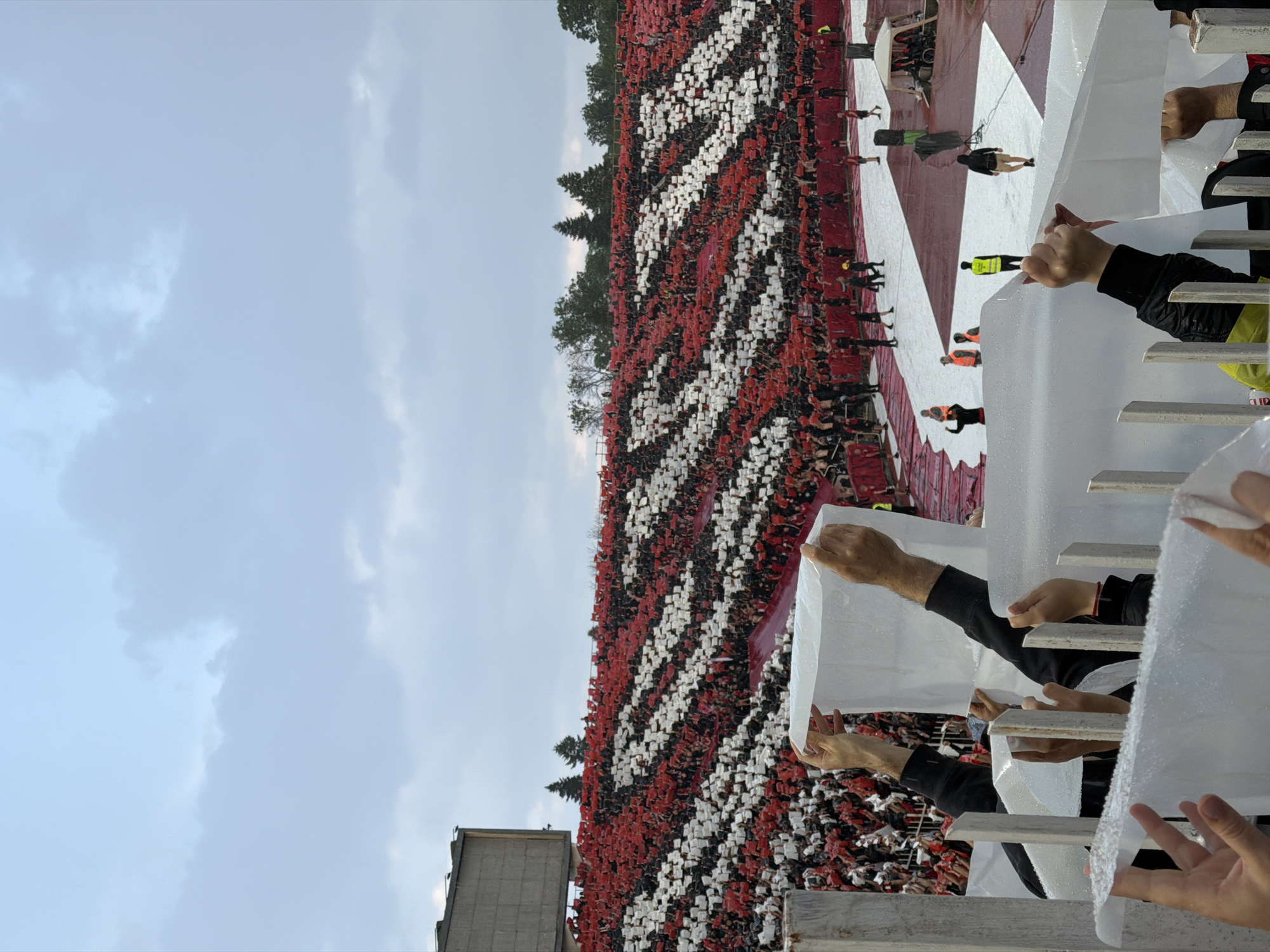 The image depicts a large crowd of people in a stadium, holding up red and white cards to create a visual display. The stadium is located in Sofia, Bulgaria, as evidenced by the architecture of the surrounding buildings and the presence of Cyrillic script in the crowd. The people appear to be attending a sporting event, possibly a football match, given the scale of the crowd and the organized display. The photo is taken from behind a barrier, with hands holding up white sheets or plastic coverings, likely for protection from the elements. The sky is overcast, suggesting a cloudy day. The overall scene conveys a sense of collective excitement and participation in a large-scale event.