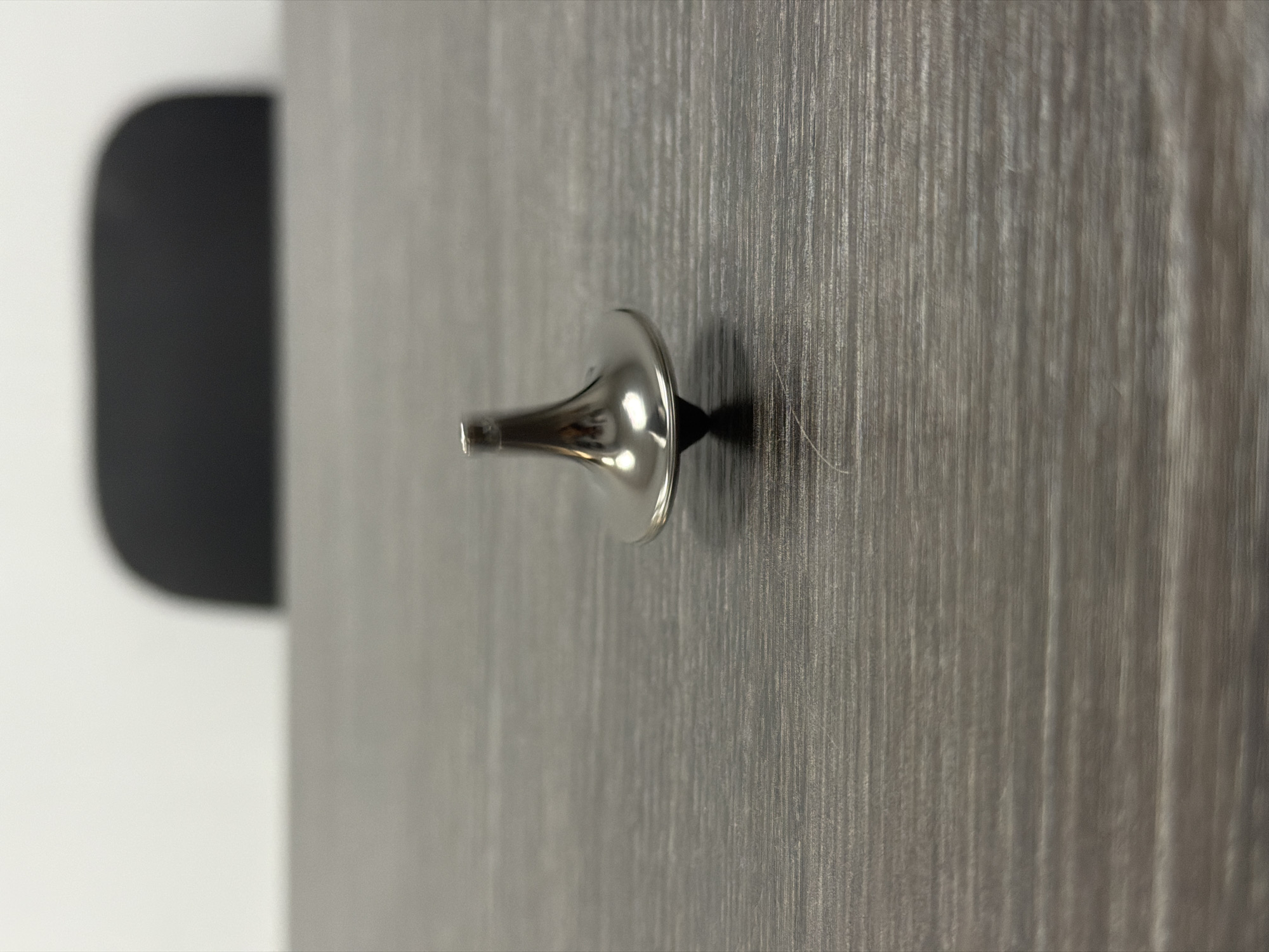 The scene depicts a polished, silver-colored metal spinning top standing upright on a dark gray, horizontally-grained laminate surface. The spinning top features a conical base, a wider, reflective mid-section, and a tapering upper shaft with a flat top. A subtle shadow is cast beneath the top's base. A single light-colored fiber is visible on the laminate surface to the lower right of the top. In the background, a large, dark, blurry object with a rounded top edge, consistent with the back of an office chair, is positioned against a uniformly light-colored wall. The environment is an indoor setting, likely an office or workspace, located in the city of Unknown, Unknown. No individuals or active interactions are present.