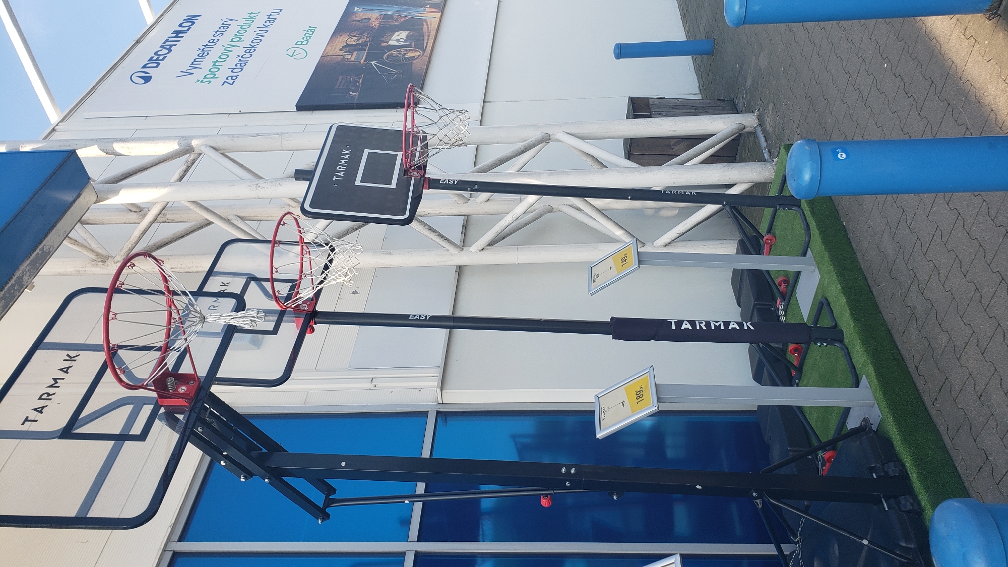 The image shows several Tarmak basketball hoops set up outdoors, likely in front of a Decathlon store in Prešov, Slovakia. The basketball hoops vary in size and are mounted on black poles. Some of the poles have price tags attached. The ground is paved with interlocking bricks and a small patch of artificial grass is visible. In the background, there is a white building with blue-tinted windows and a large banner with the Decathlon logo and text in Slovak. The scene appears to be a retail display for sporting goods. There are no people visible in the image.