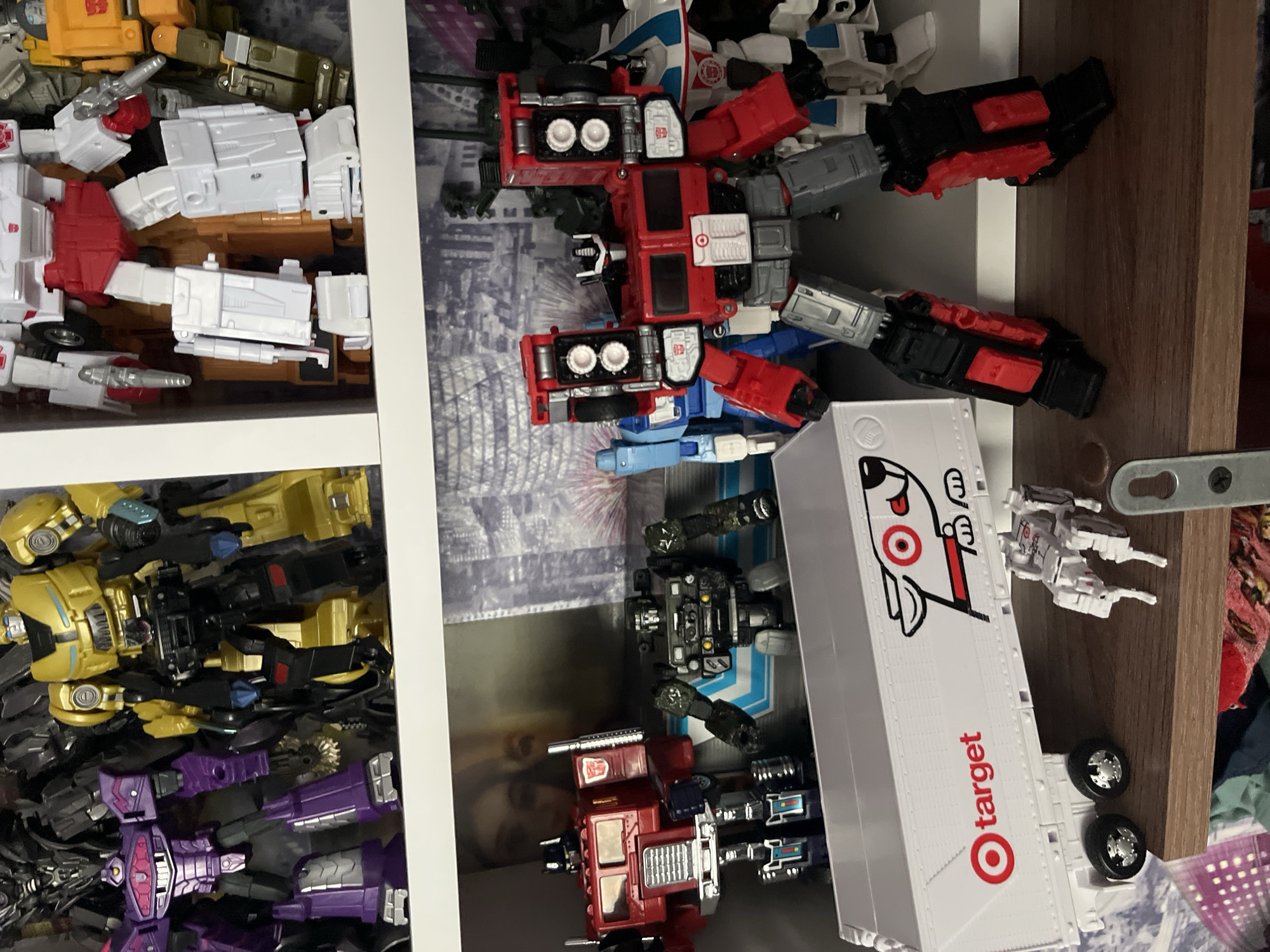 The image shows a shelf filled with what appear to be Transformers toys. The toys are mostly in robot or vehicle mode and are arranged haphazardly in the compartments of the shelf. One toy is a white truck trailer with the Target logo on its side. The background shows a photograph of what looks like a cityscape, blurred and partially obscured by the toys. The image was not taken in Марковский, Russia, because the text on the truck is in English.